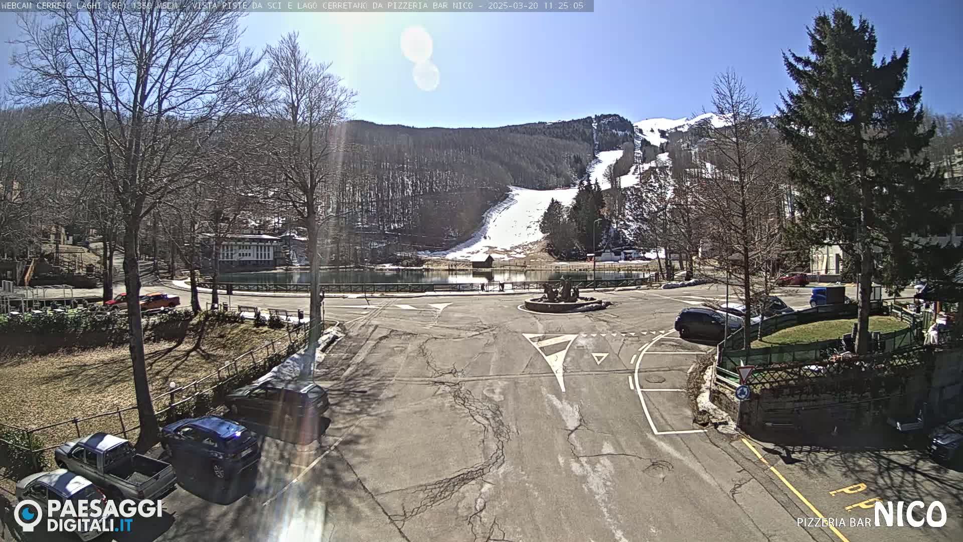 A sunny day reveals a parking lot overlooking a snow-covered ski slope and a calm lake, surrounded by trees and buildings.