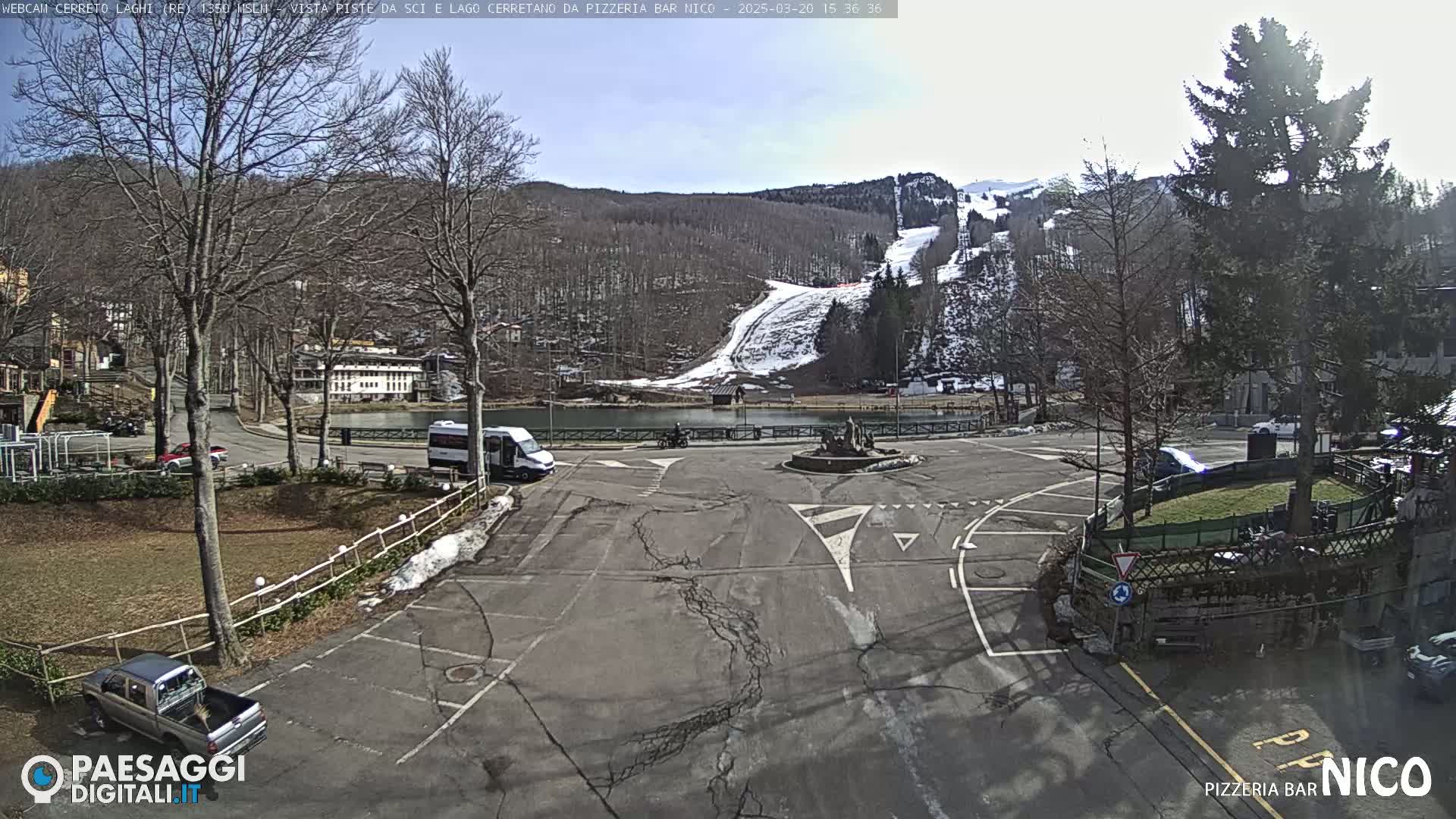 A sunny day reveals a parking lot with a few vehicles, a small lake, and a snow-covered ski slope in the background.