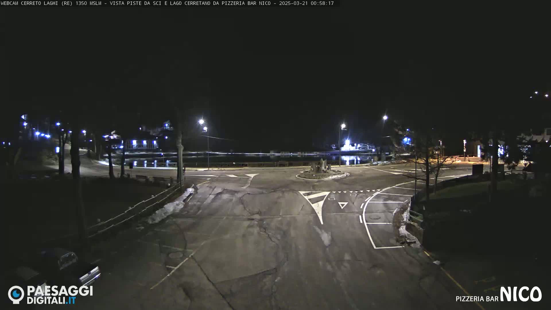 A nighttime view of a mostly empty roundabout and surrounding roads, with some snow on the ground and a calm body of water visible in the background.