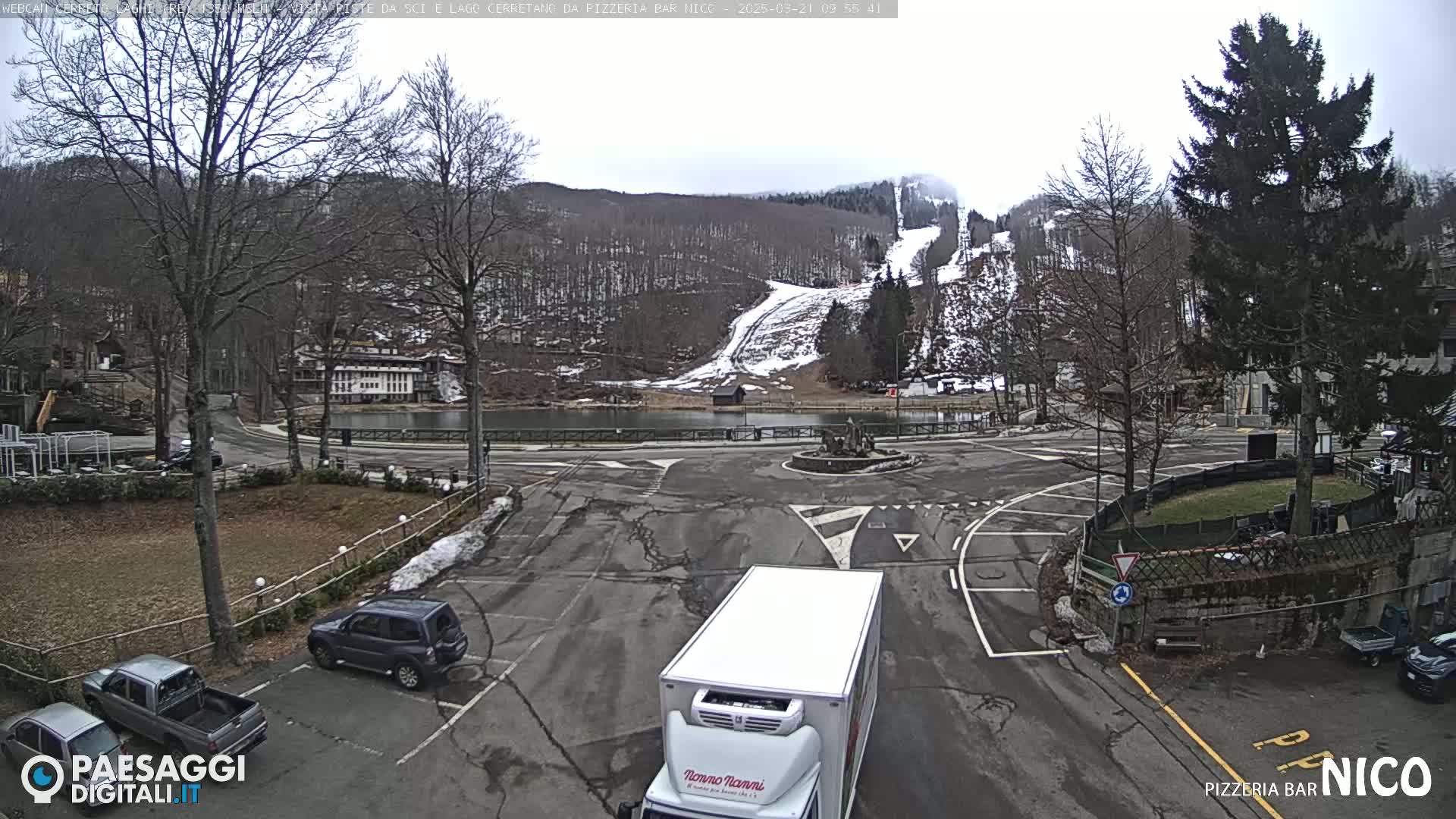 A parking lot with several vehicles is situated near a body of water and a snow-covered ski slope in the background under an overcast sky.