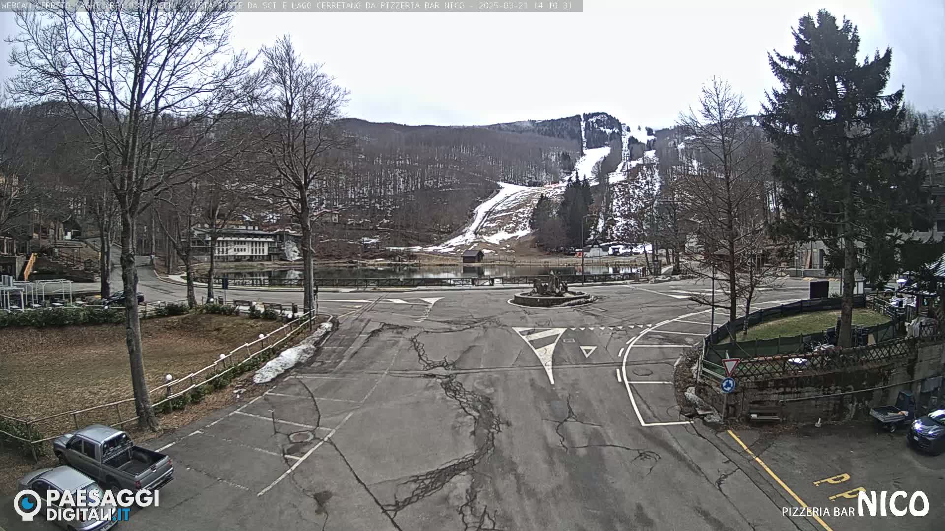 A mostly empty parking lot and roundabout are situated near a small lake and ski slopes, under an overcast sky.