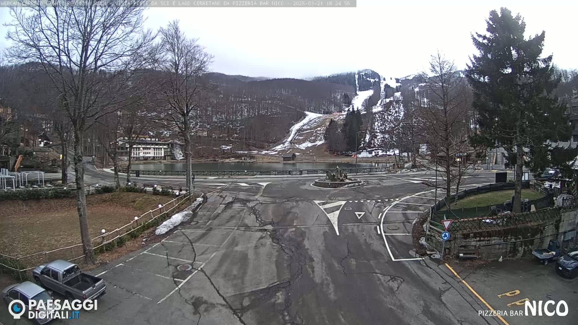 An overcast day reveals a parking lot and roadway curving around a small lake, with a ski slope visible on a snow-covered hillside in the background.