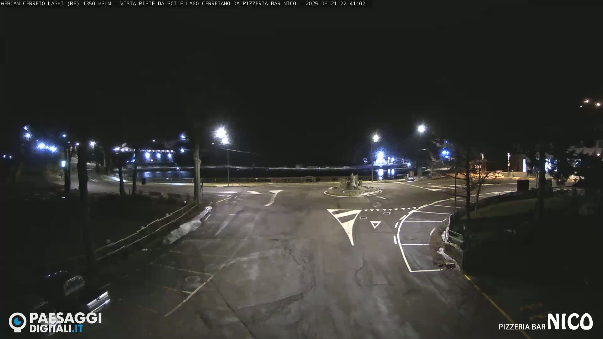 A nighttime view of a mostly empty parking lot and roadway near a body of water, illuminated by streetlights.