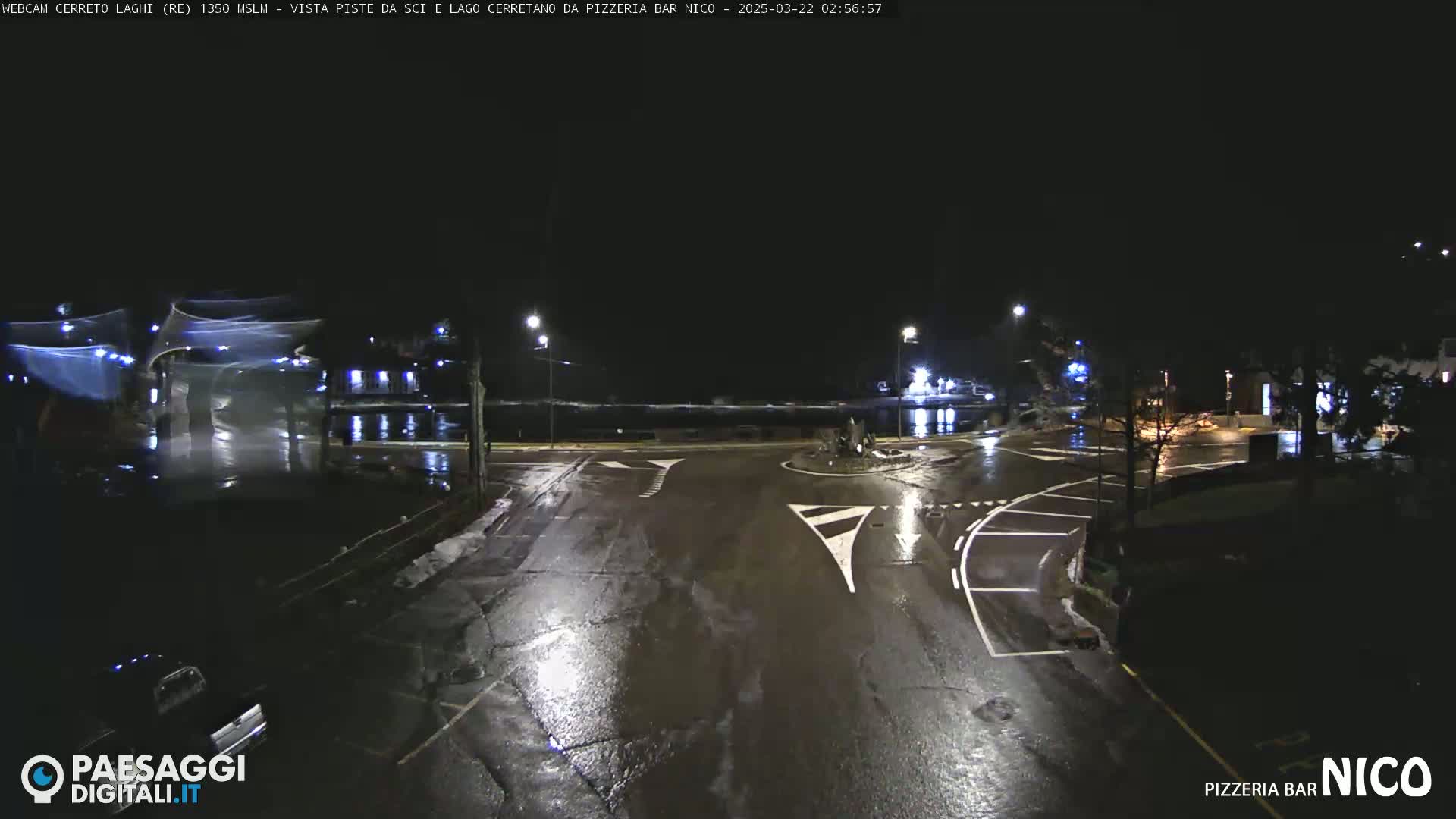 A wet, nighttime view of a mostly empty intersection with streetlights and a few parked cars.