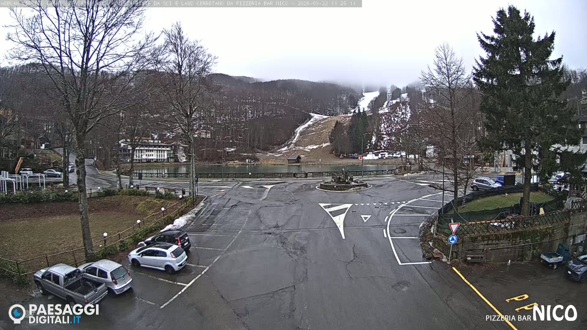 A parking lot with several cars is situated near a small lake, with a ski slope visible in the background under an overcast sky.