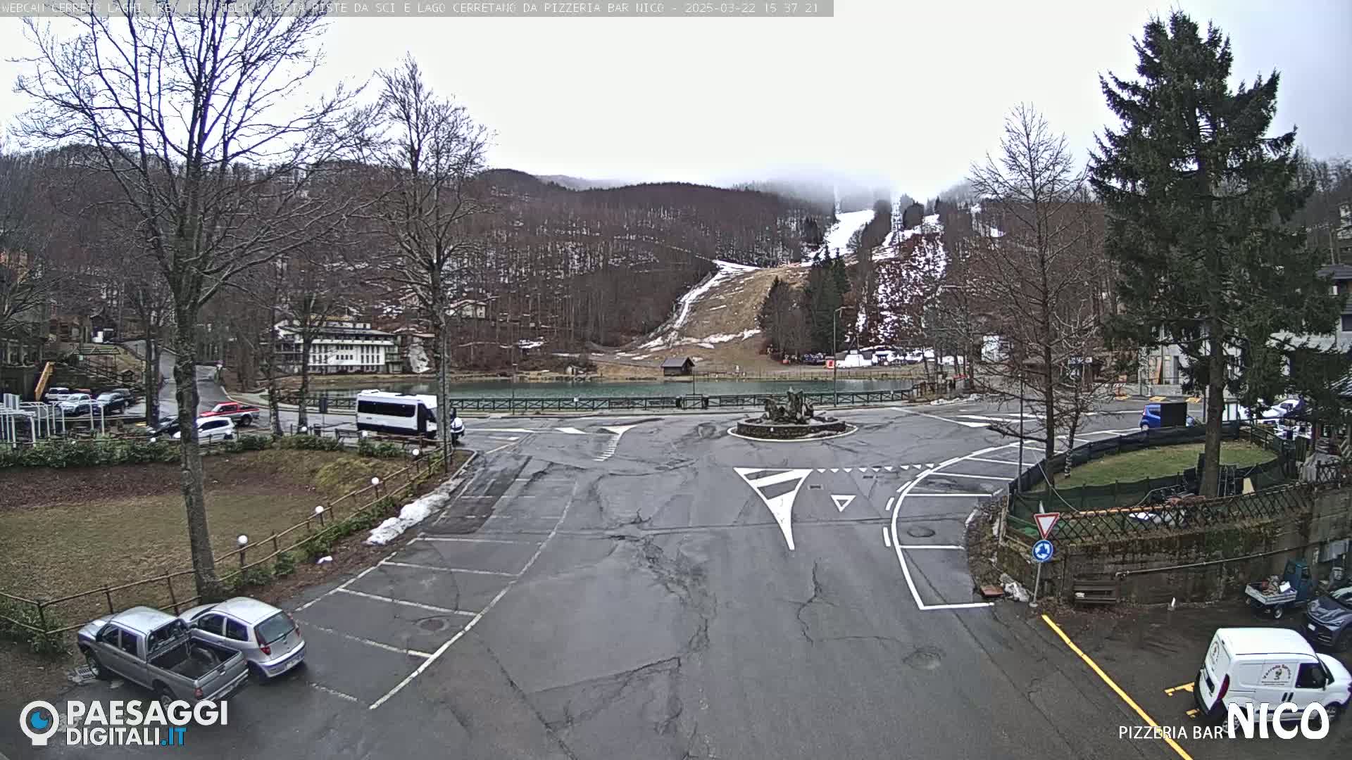 A parking lot and roadway intersect near a small lake and a wooded hillside with a ski slope visible in the overcast weather.
