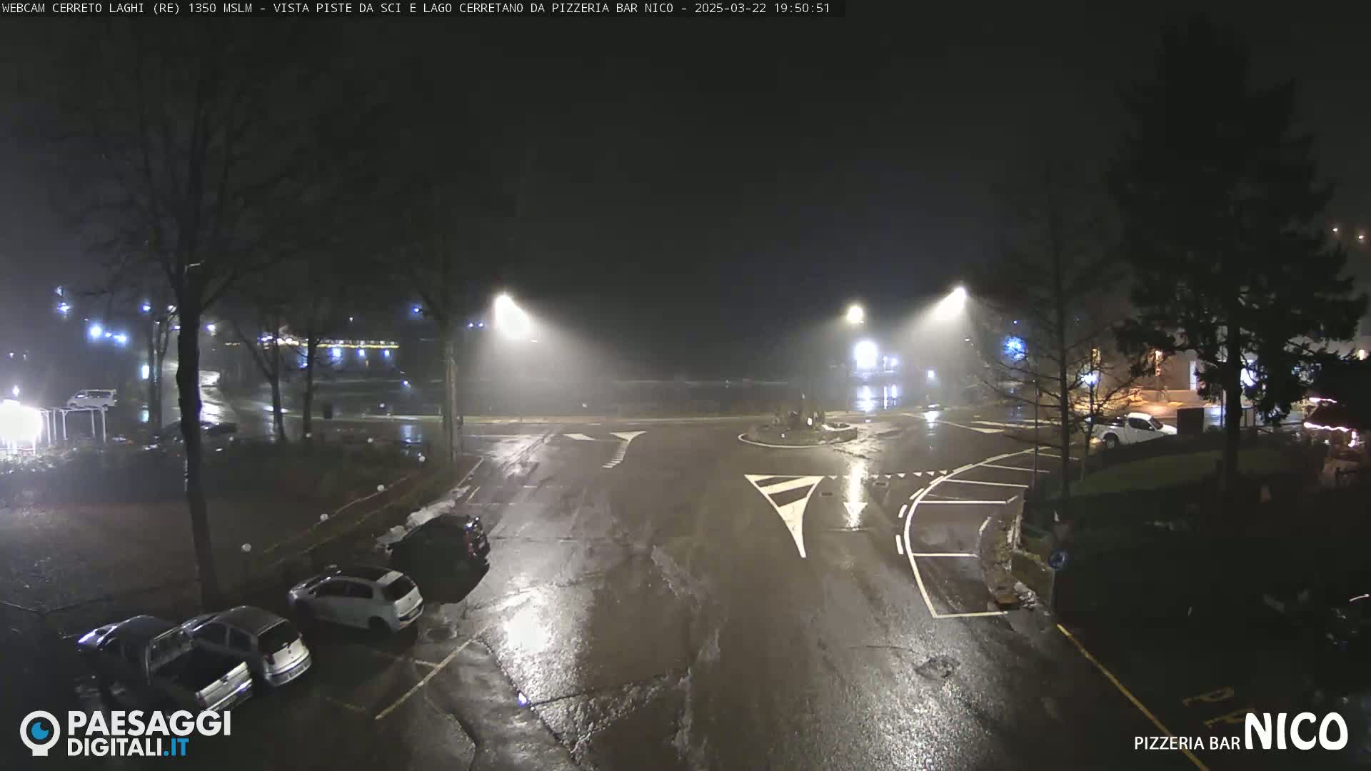 A wet, nighttime scene shows a mostly empty parking lot and a roundabout with several cars parked nearby, illuminated by streetlights in foggy conditions.