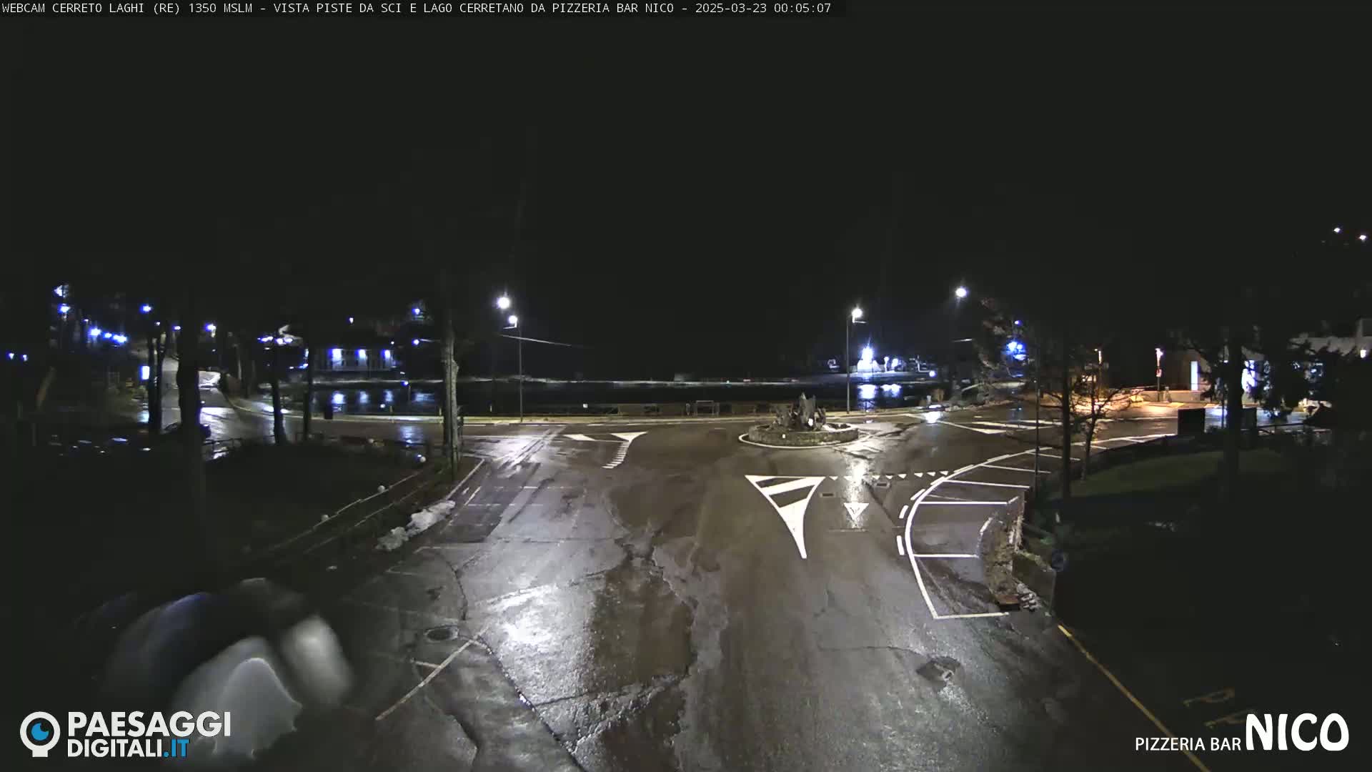 A wet, dark nighttime scene shows an empty intersection with streetlights and a body of water in the distance.