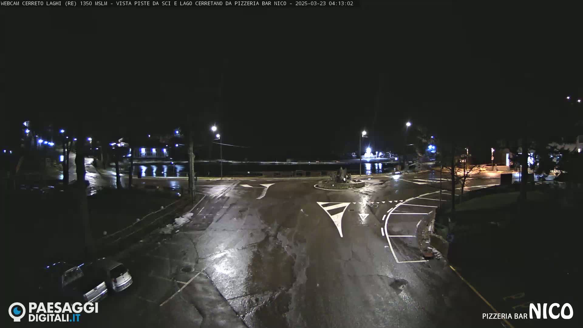 A wet, nighttime street scene shows a roundabout and parking lot with a few cars, illuminated by streetlights.
