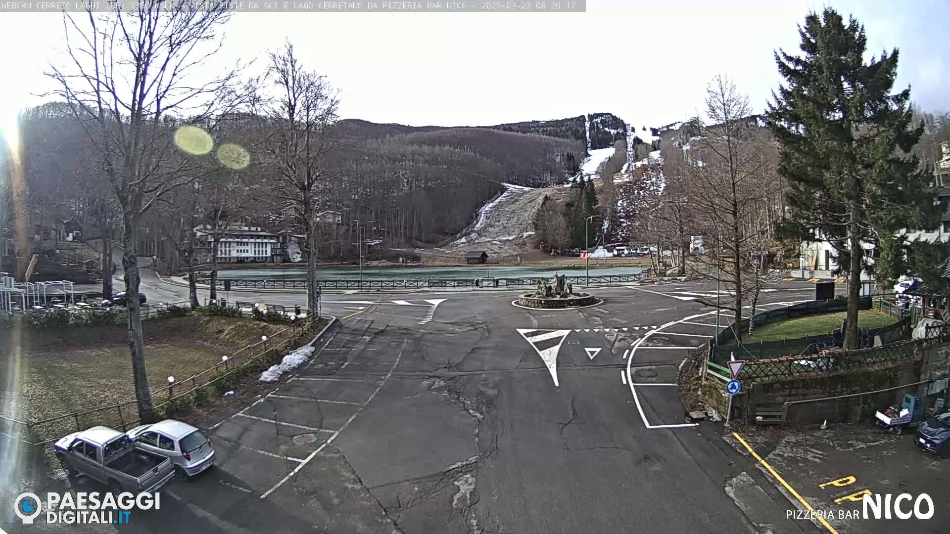 A parking lot with a few cars, situated near a roundabout and a partially snow-covered ski slope, on a partly sunny day.