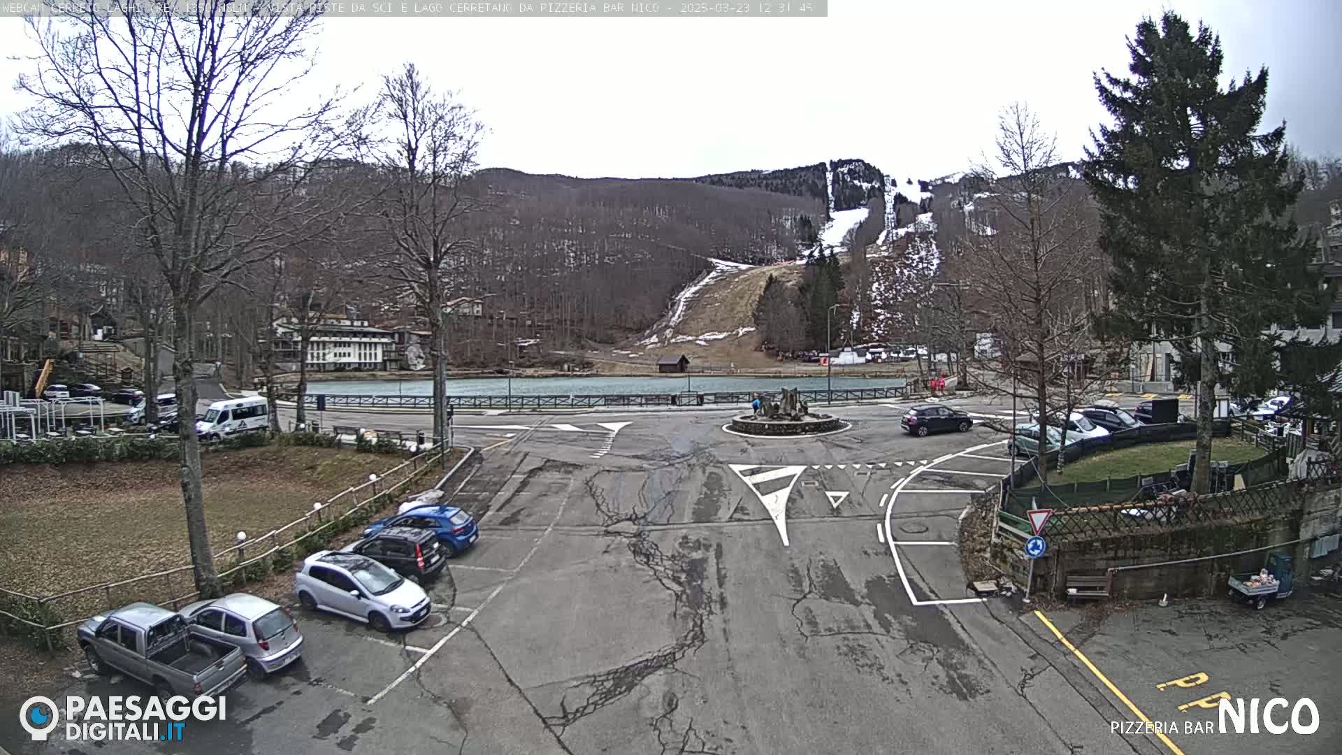 A parking lot and roundabout are situated in front of a body of water and a ski slope on a cloudy day, surrounded by bare deciduous trees and evergreen conifers.
