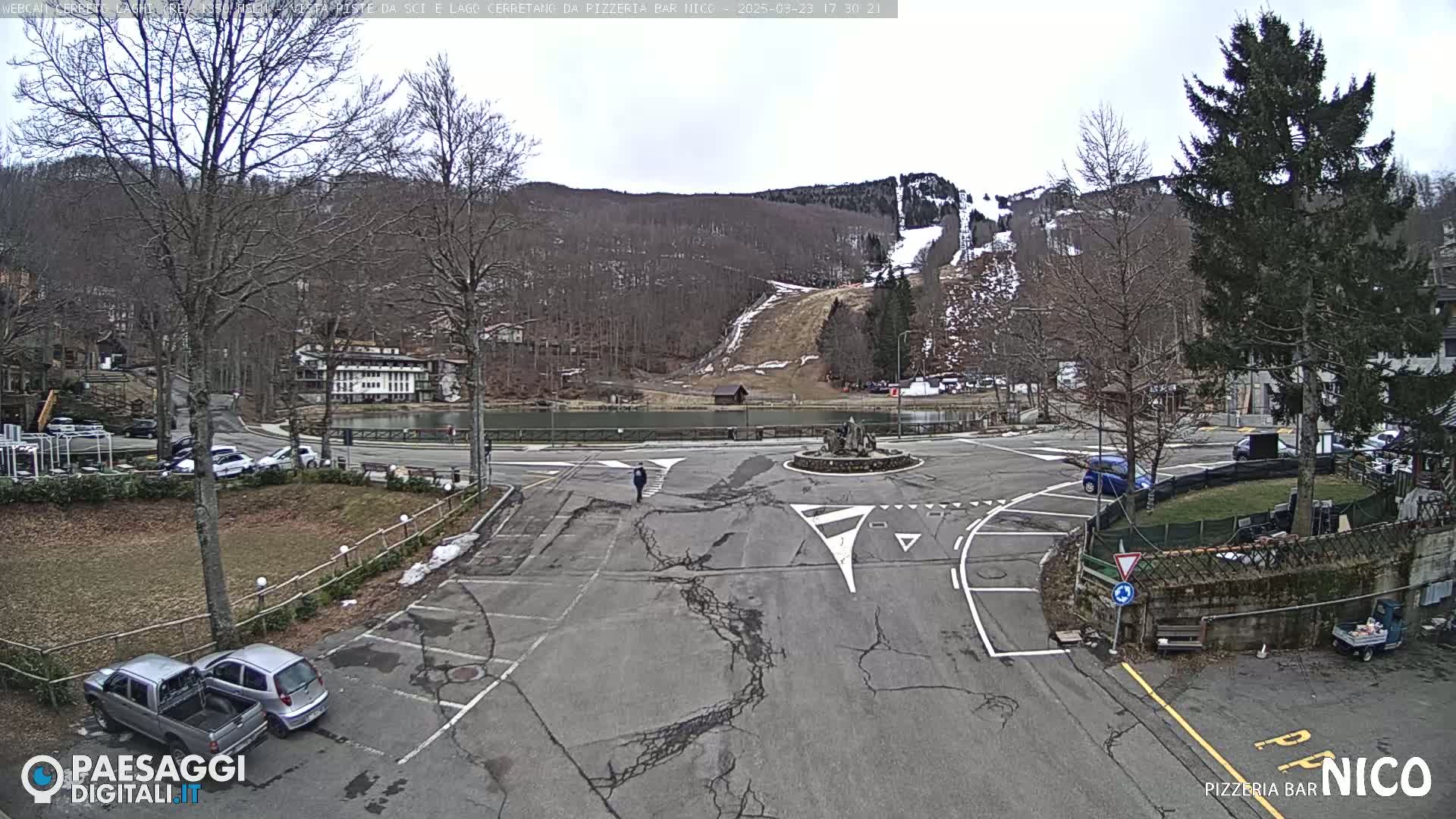 A mostly empty parking lot and roundabout are situated in front of a small lake and a ski slope on a cloudy day.