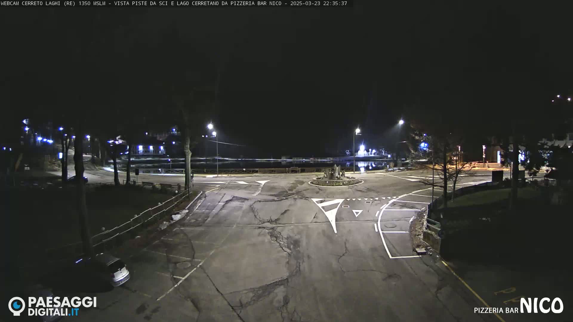 A nighttime, overhead view of an empty parking lot and roadway intersection, illuminated by streetlights.