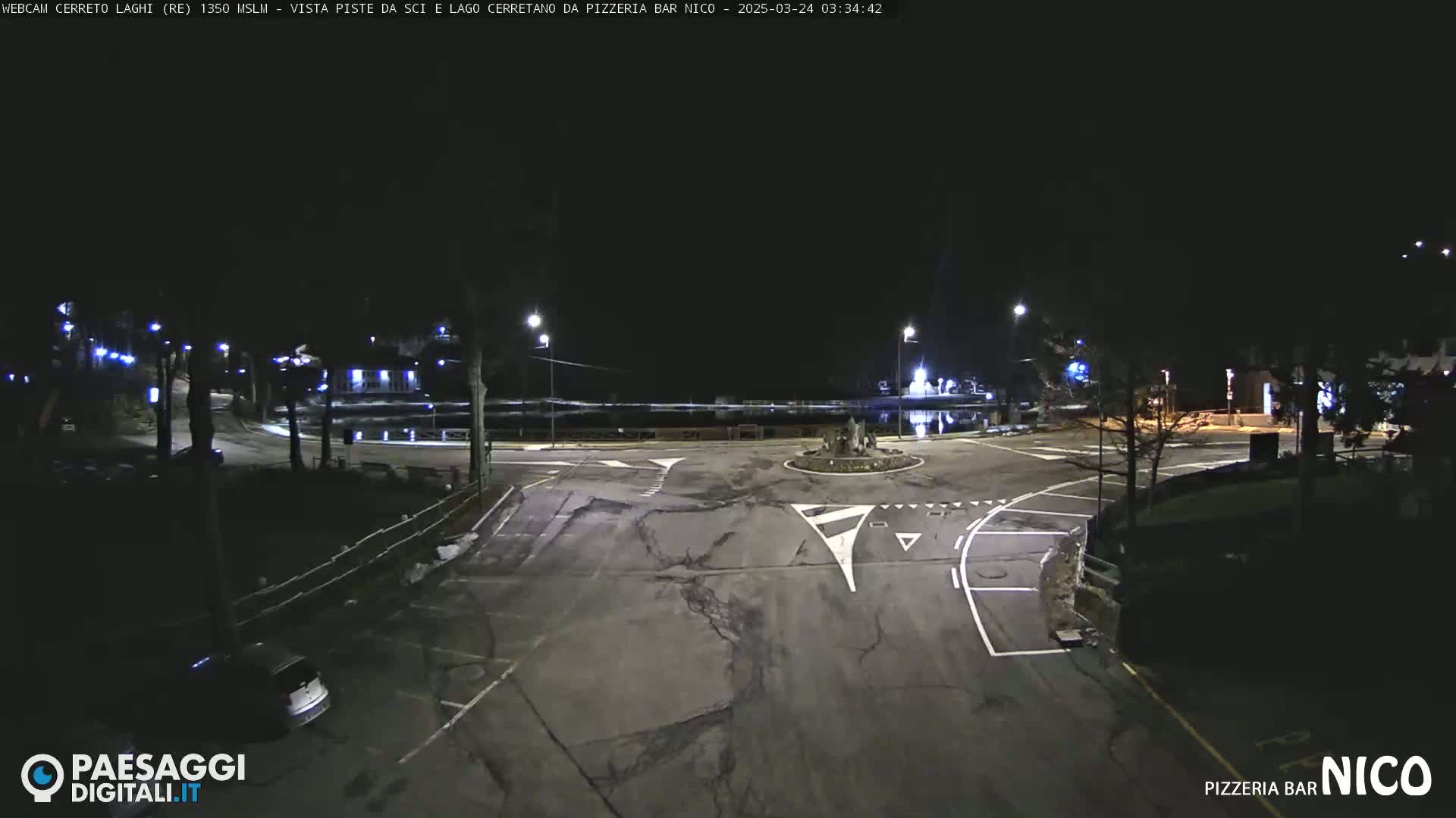 A nighttime view of a mostly empty parking lot and roundabout near a body of water, illuminated by streetlights.