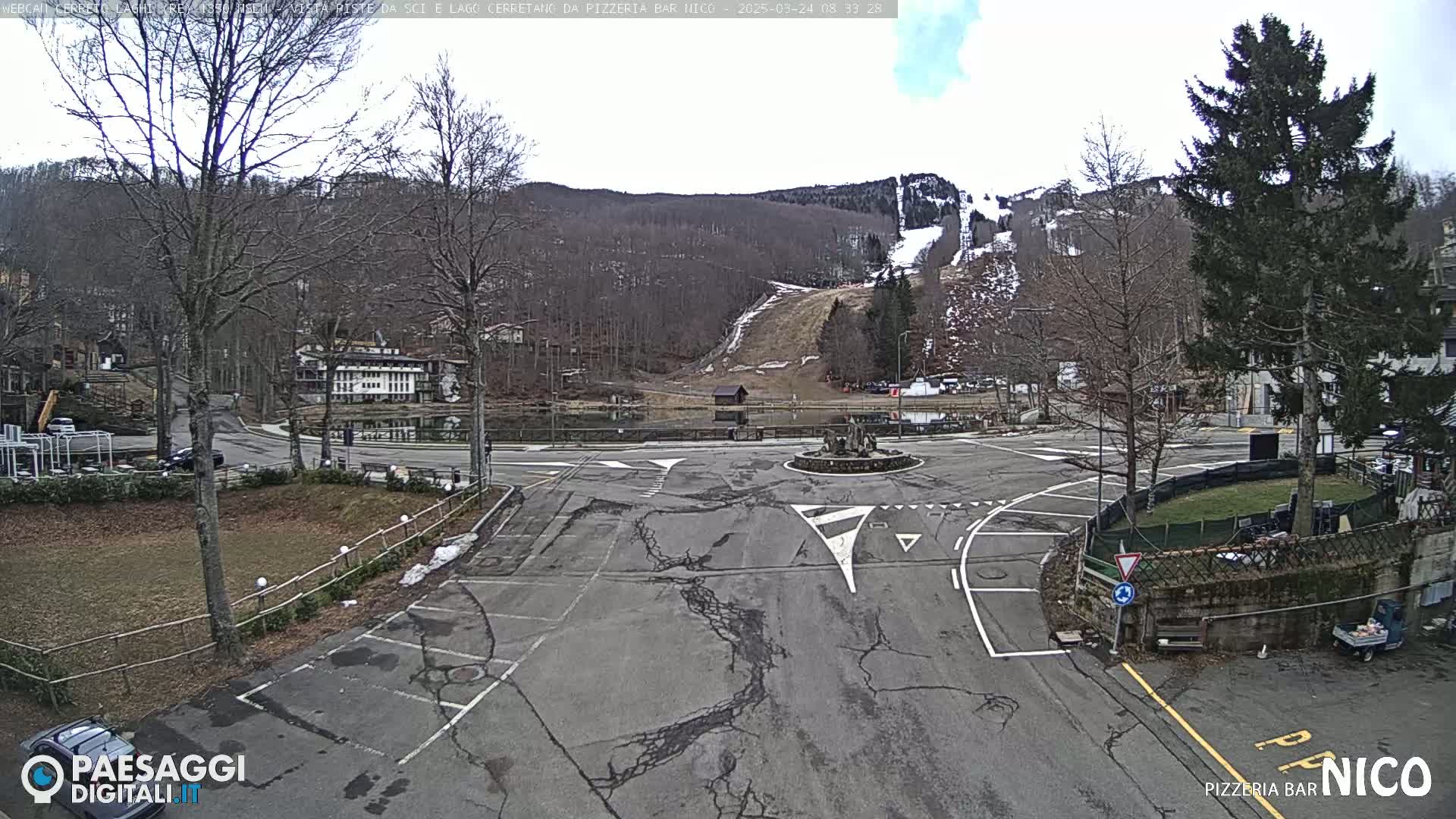 A mostly empty parking lot and roundabout surround a small pond, with a ski slope visible on a hillside in the background under a mostly cloudy sky.