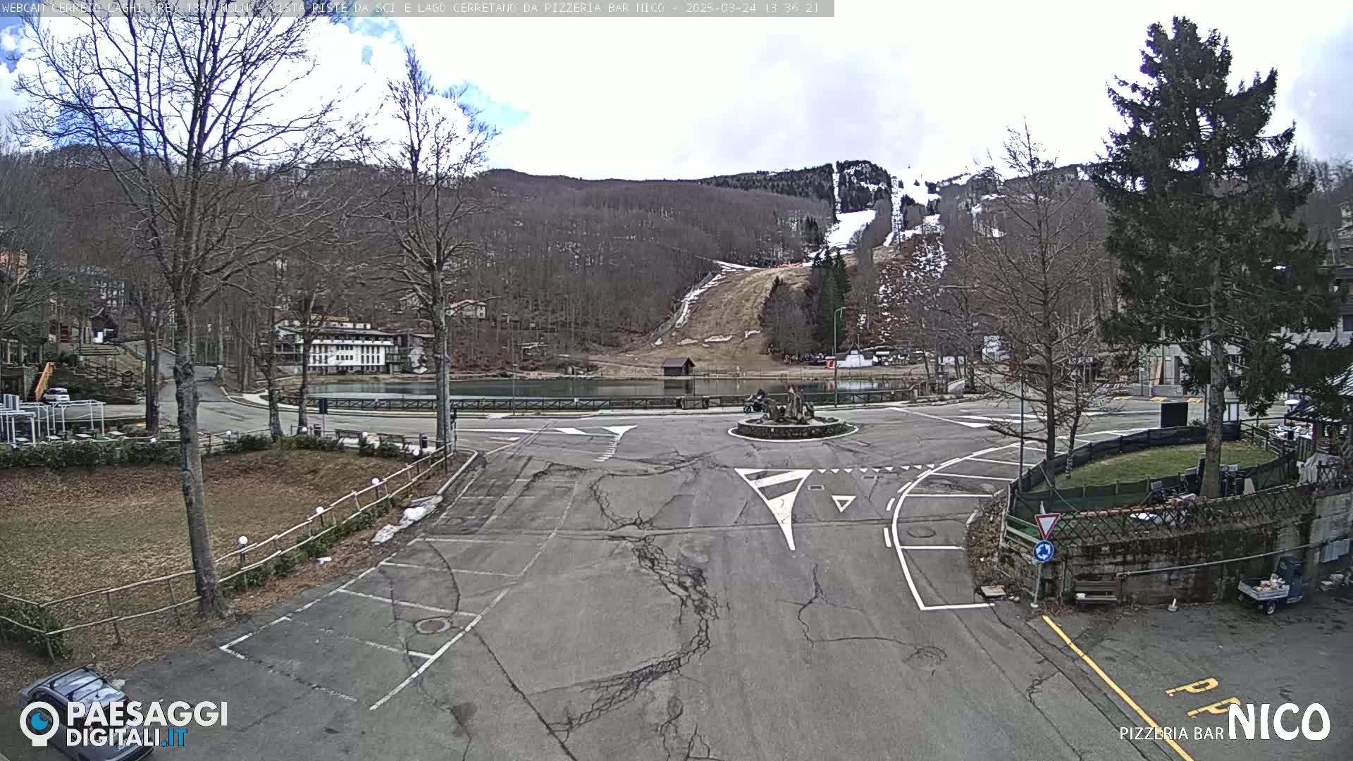 A mostly empty parking lot surrounds a roundabout in front of a small lake, with a ski slope visible on a hillside in the background under a partly cloudy sky.