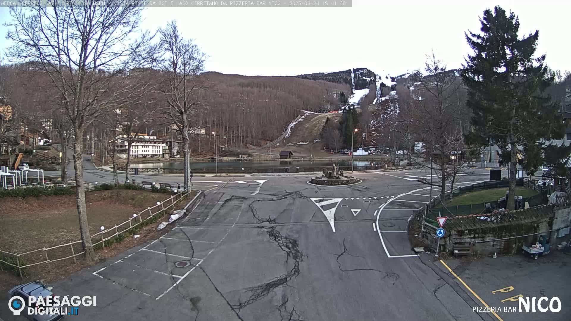 A mostly empty parking lot surrounds a roundabout in front of a lake and ski slope under an overcast sky.