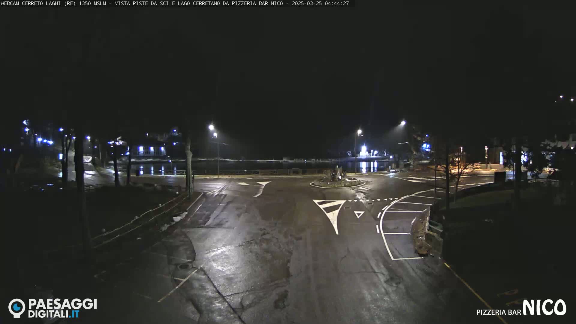 A wet, empty asphalt roundabout and roads are illuminated at night by streetlights, with a body of water visible in the background.