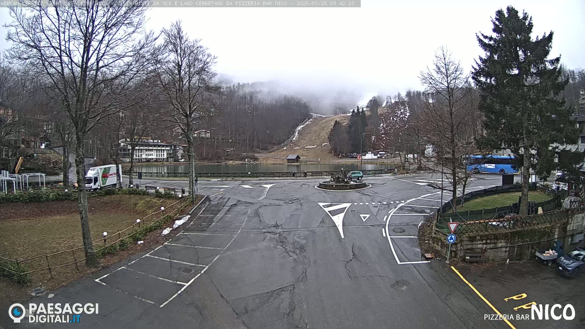 A mostly empty parking lot with a roundabout and a small lake is seen, under an overcast sky; a ski slope is visible in the background.
