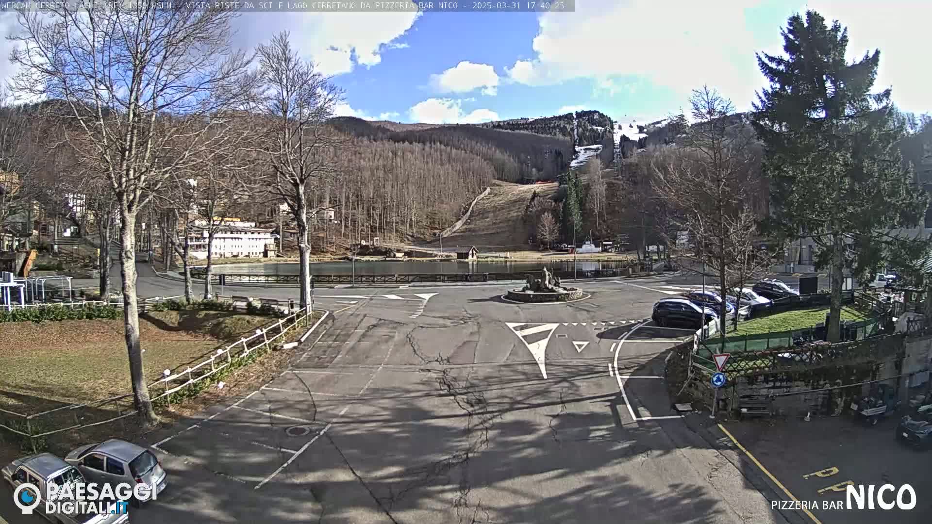 A sunny day reveals a parking lot and roundabout near a lake, with a ski slope visible on a hillside in the background.