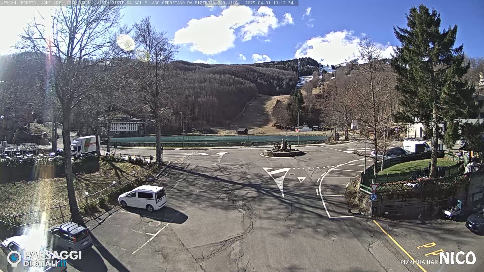 A sunny day reveals a parking lot and roundabout near a ski slope and lake, with several cars parked and some trees nearby.