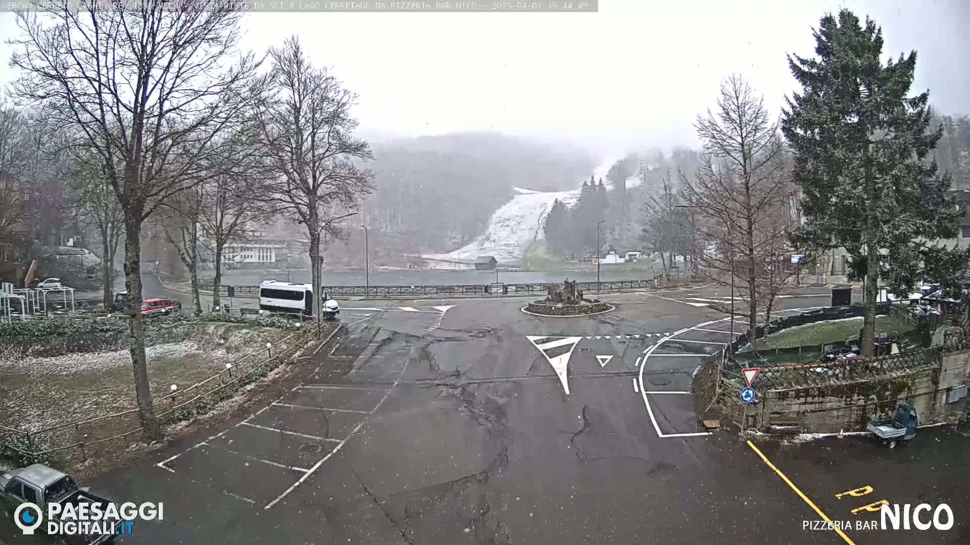 A lightly snow-covered parking lot and roadway intersect near a roundabout with a ski slope visible in the background under a light snowfall.