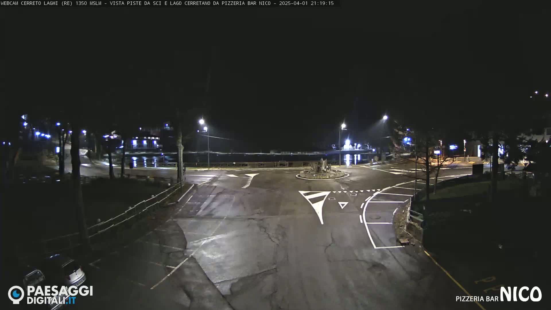 A wet, empty parking lot and roundabout are illuminated at night by streetlights.
