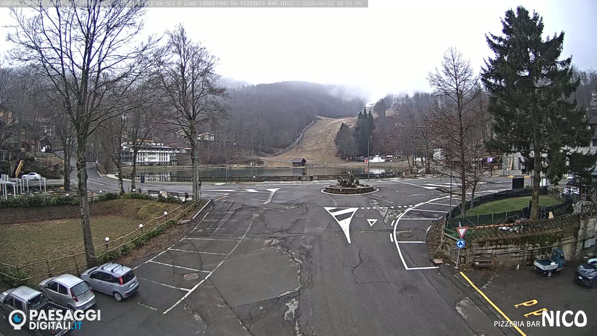 A parking lot and roundabout are seen, with a misty mountain in the background and several cars parked.