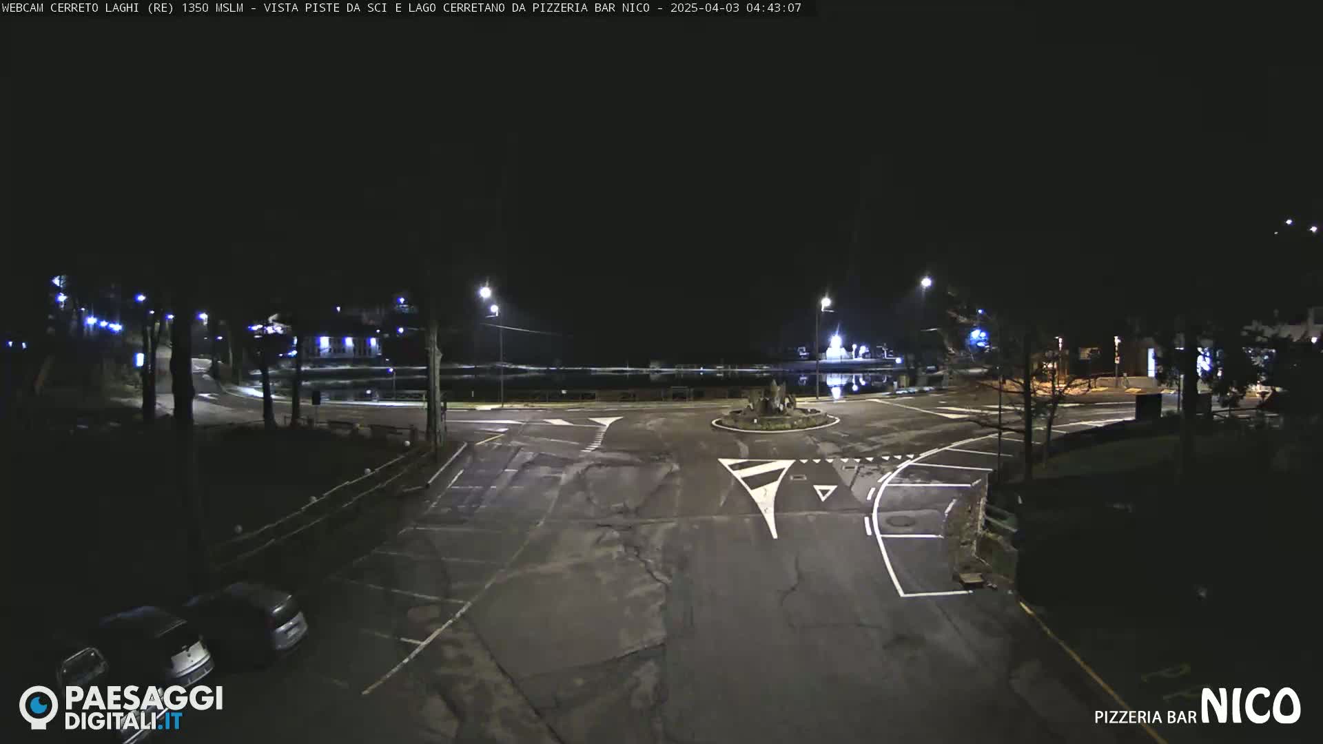A dark, nighttime view of a mostly empty roundabout and parking lot, illuminated by streetlights.