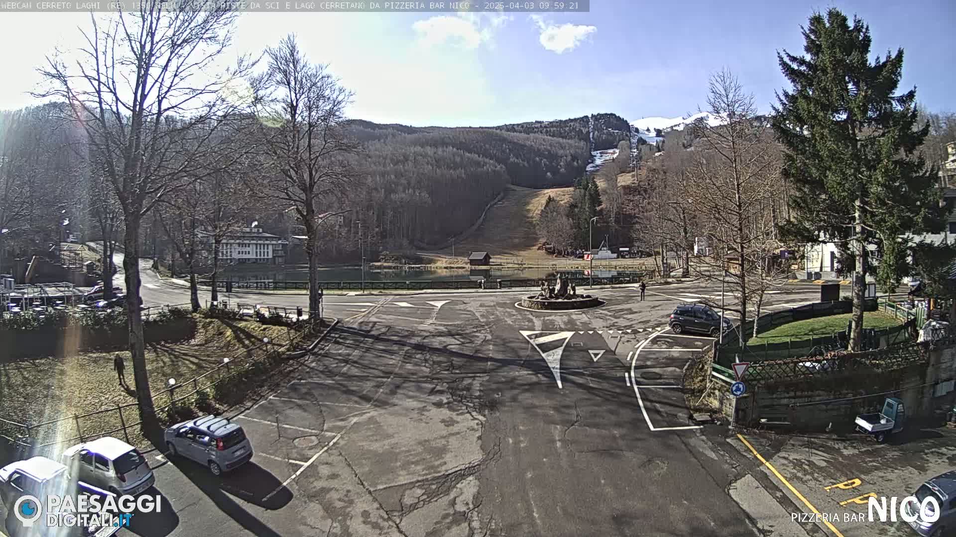 A sunny day reveals a parking lot and roundabout near a lake and ski slope in a mountainous area.