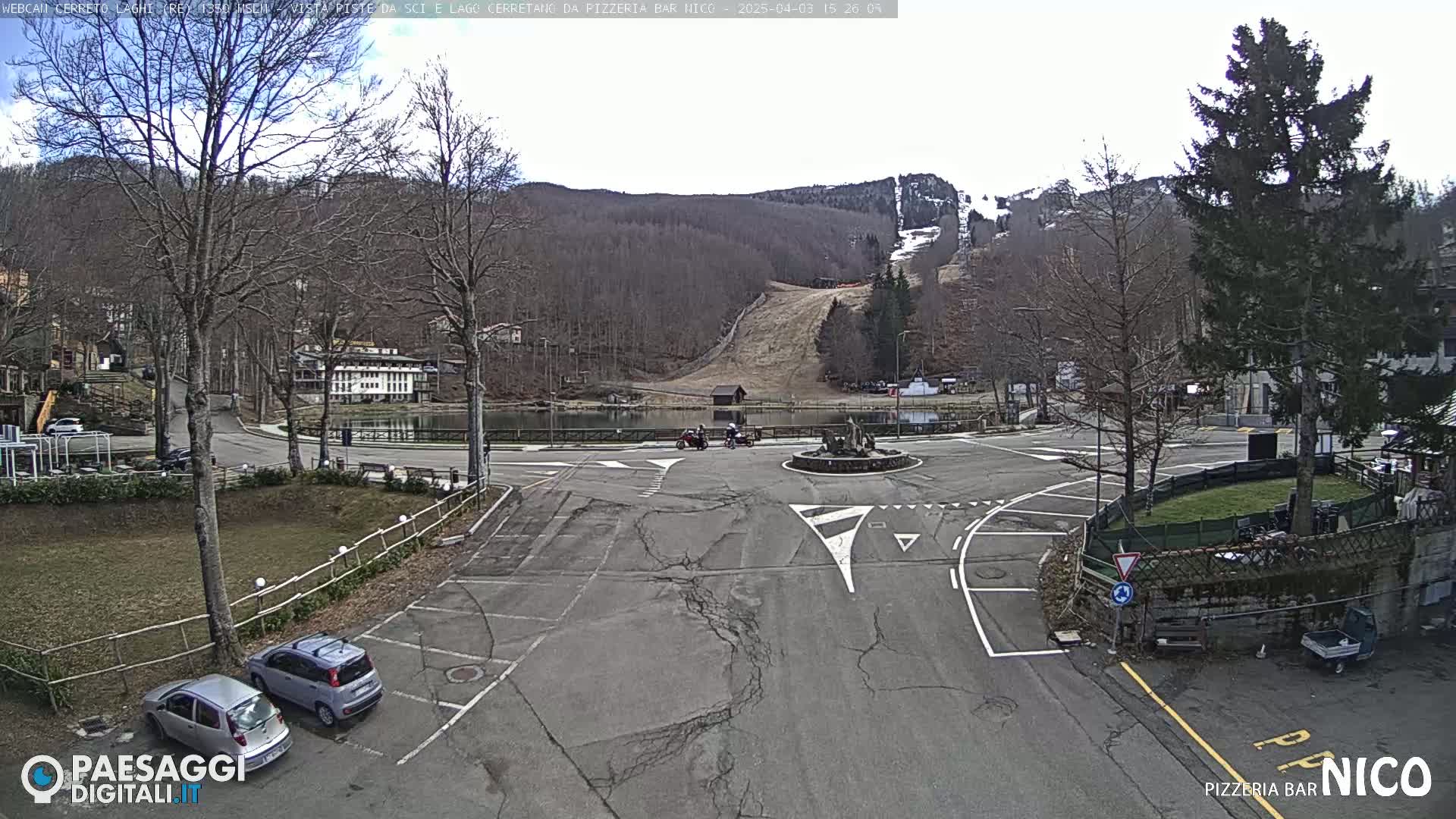 A mostly empty parking lot and roundabout overlook a small lake and a ski slope on a partly cloudy day.
