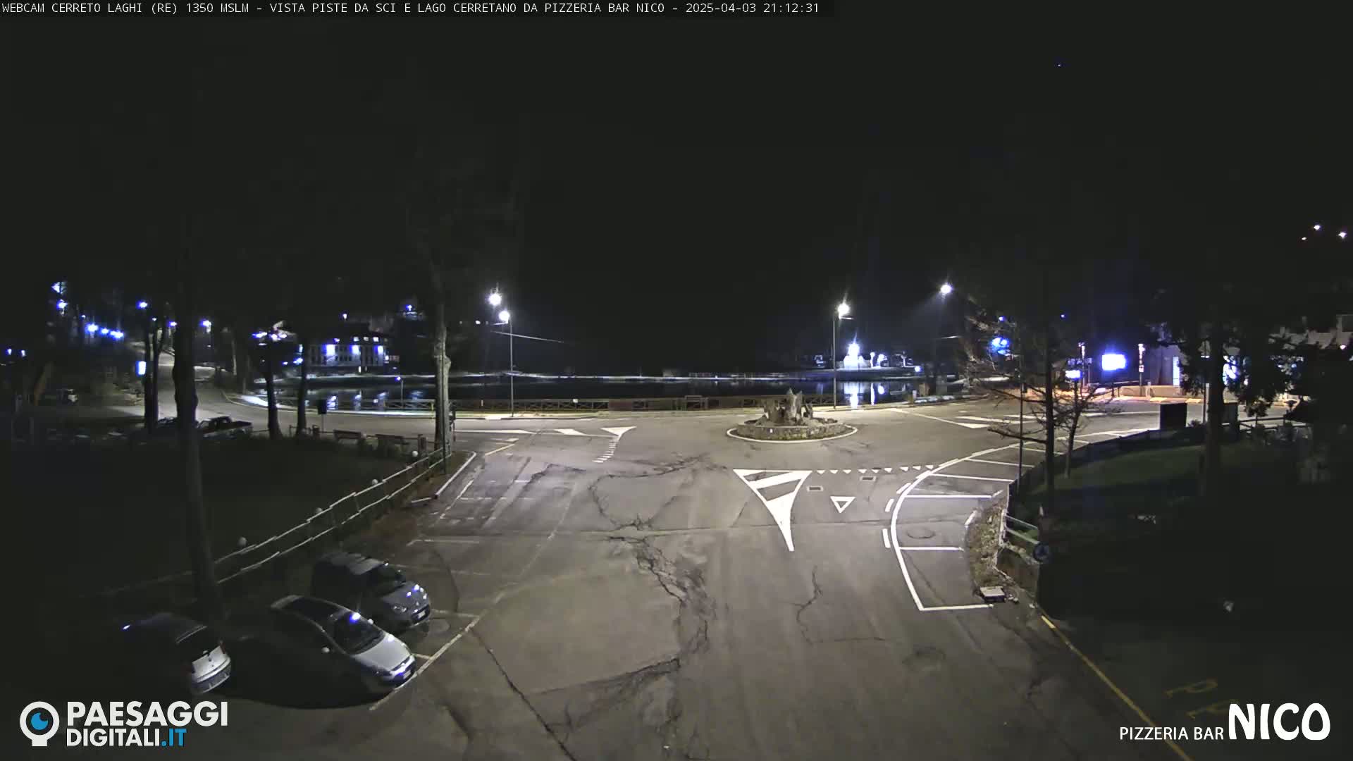 A nighttime view of a dark, empty roundabout with a few parked cars and streetlights illuminating the scene.