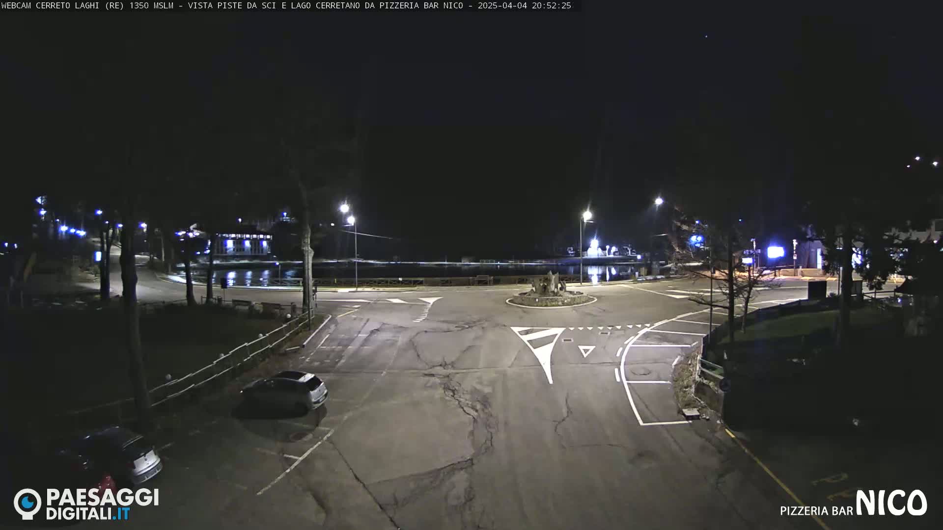 A nighttime view of a mostly empty roundabout and parking lot near a body of water, illuminated by streetlights.