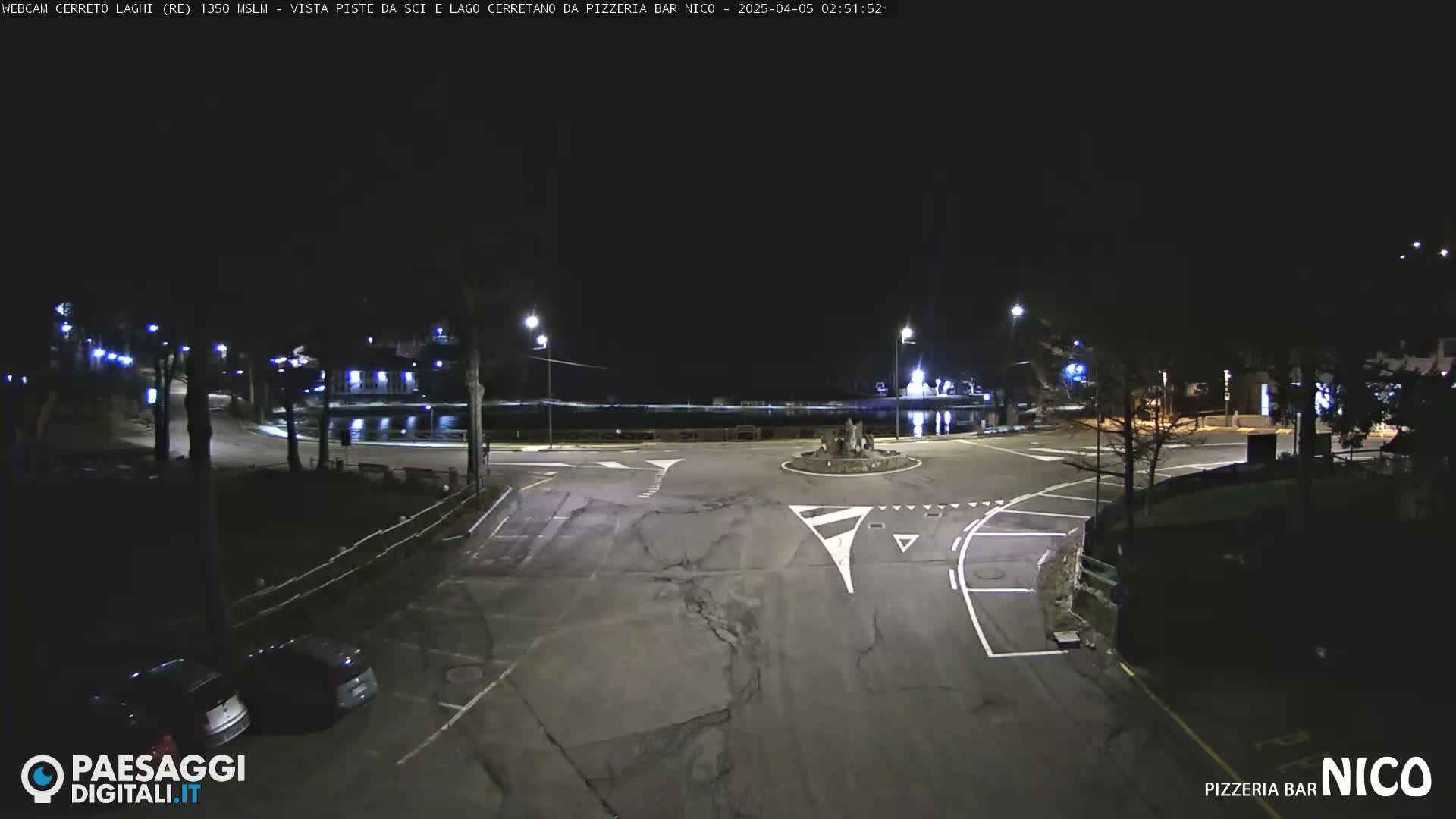 A nighttime view of a dark, empty parking lot and roadway intersection near a body of water, illuminated by streetlights.