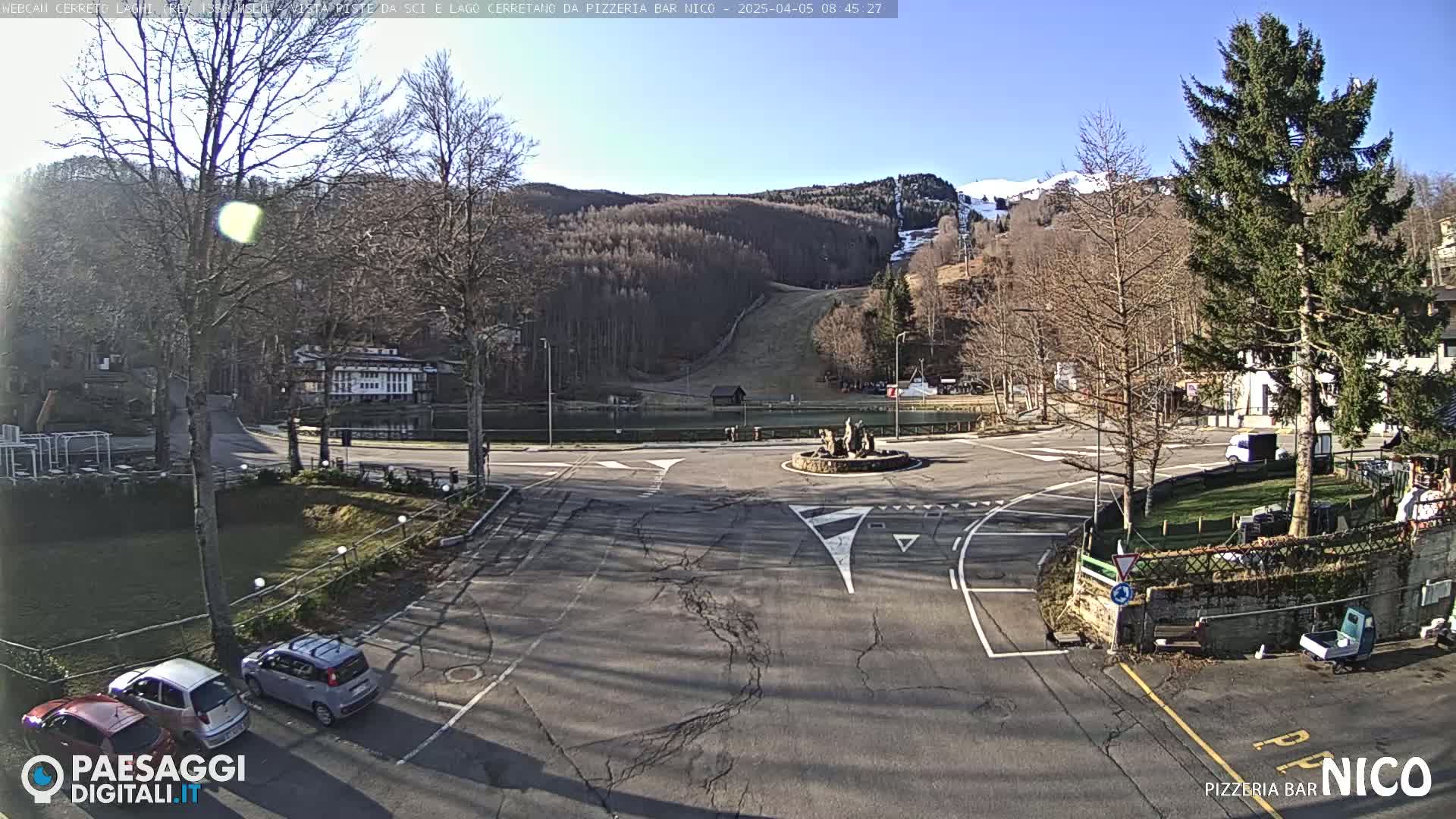 A sunny day shows a paved roundabout with several cars parked nearby, situated in front of a small town nestled at the base of a snow-dusted mountain with ski slopes visible in the background.