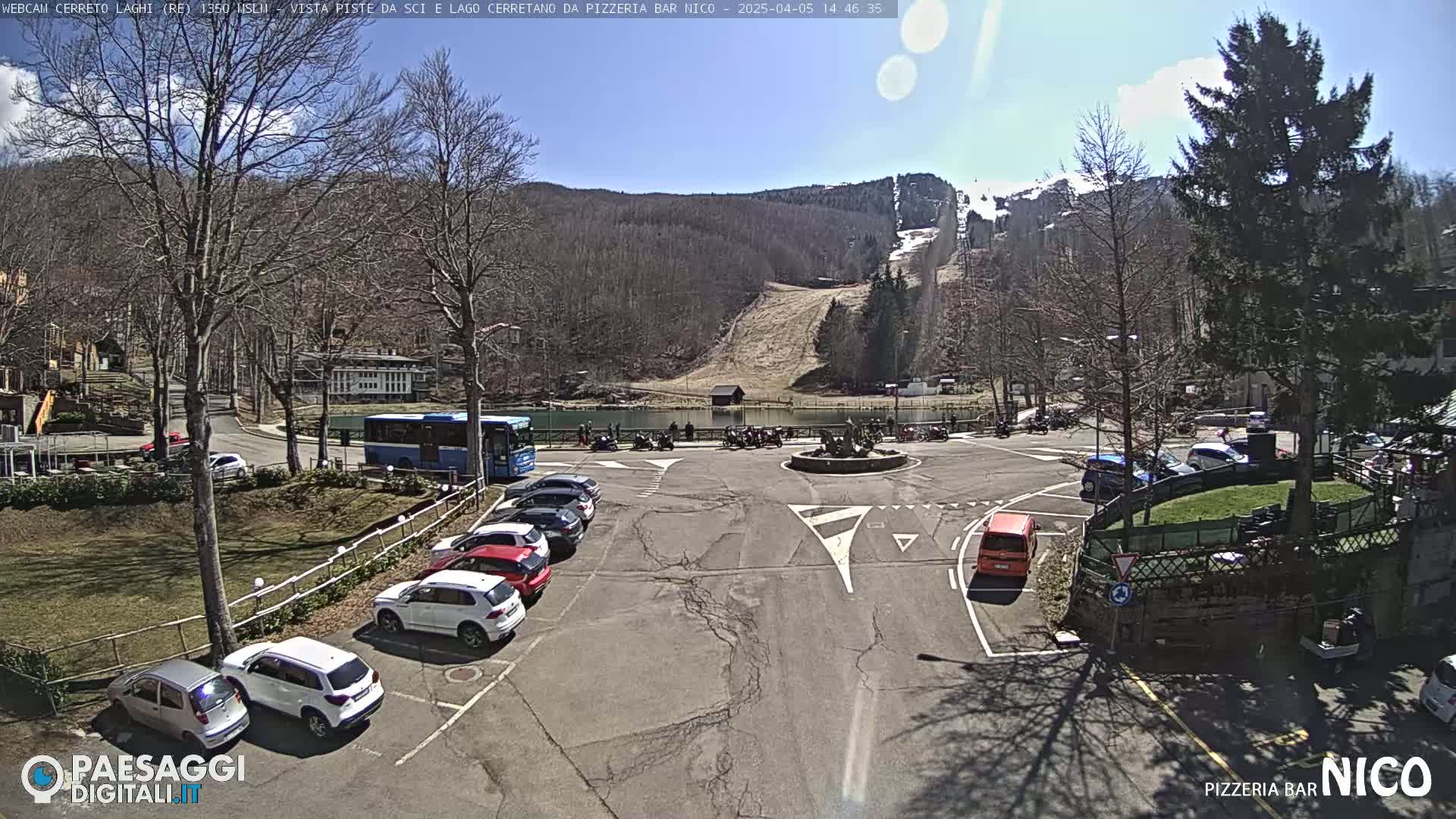 A sunny day shows a parking lot with several cars, a bus, and a ski slope in the background.