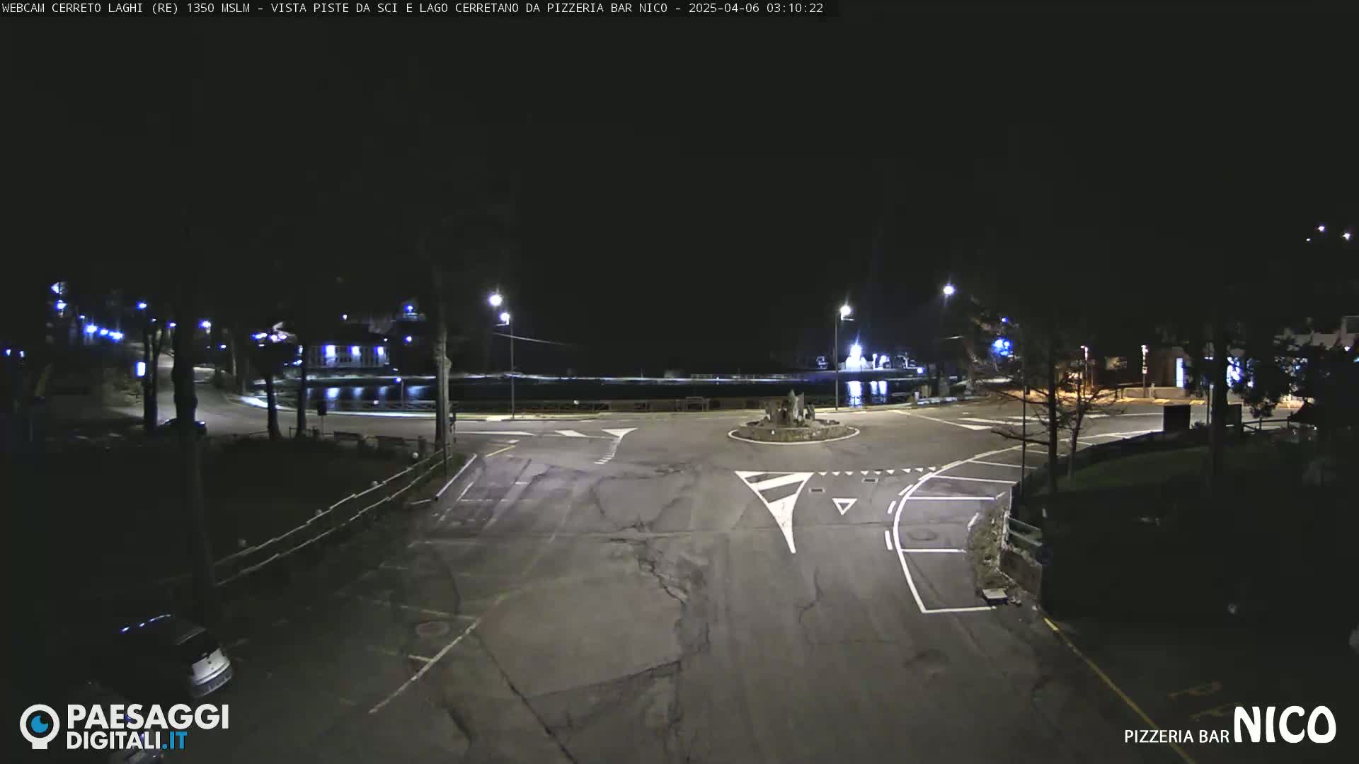 A dark, empty roundabout at night, illuminated by streetlights, is surrounded by a parking lot and sparse vegetation.