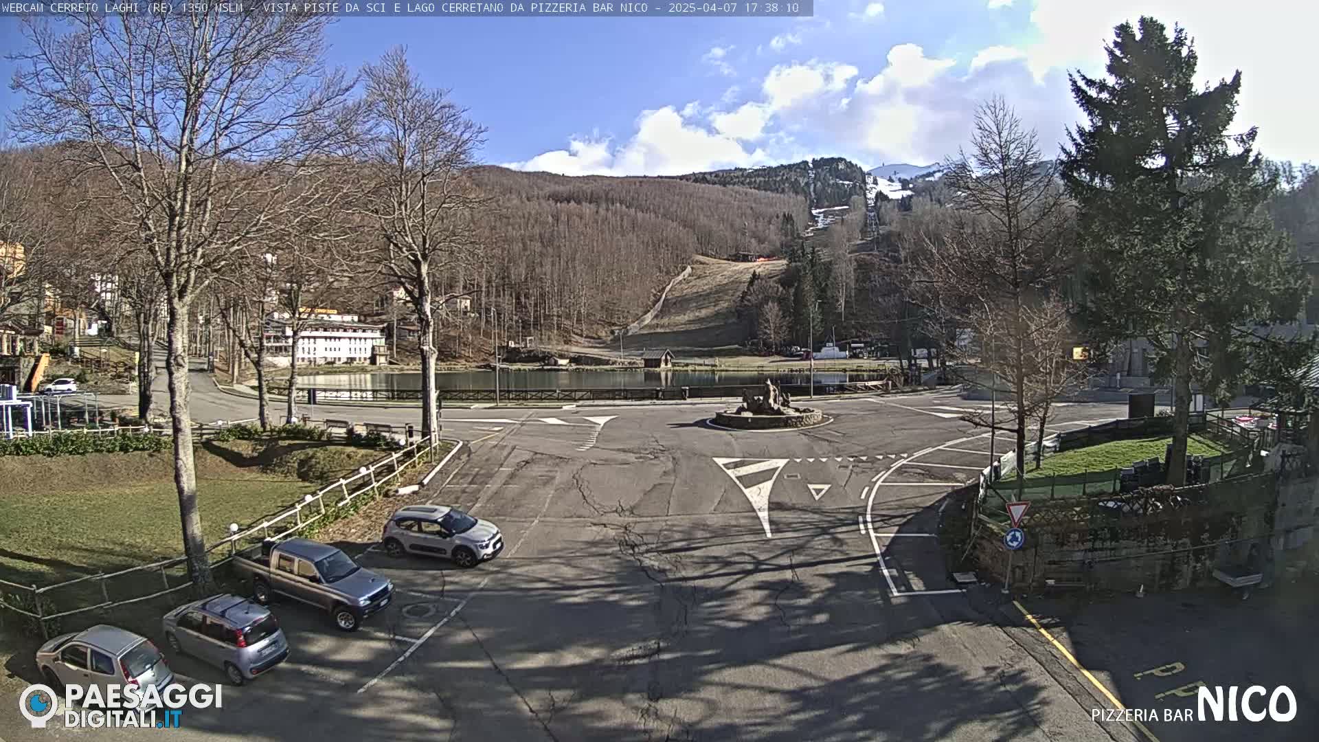 A mostly sunny day reveals a parking lot and roundabout near a lake and ski slopes in a wooded area.