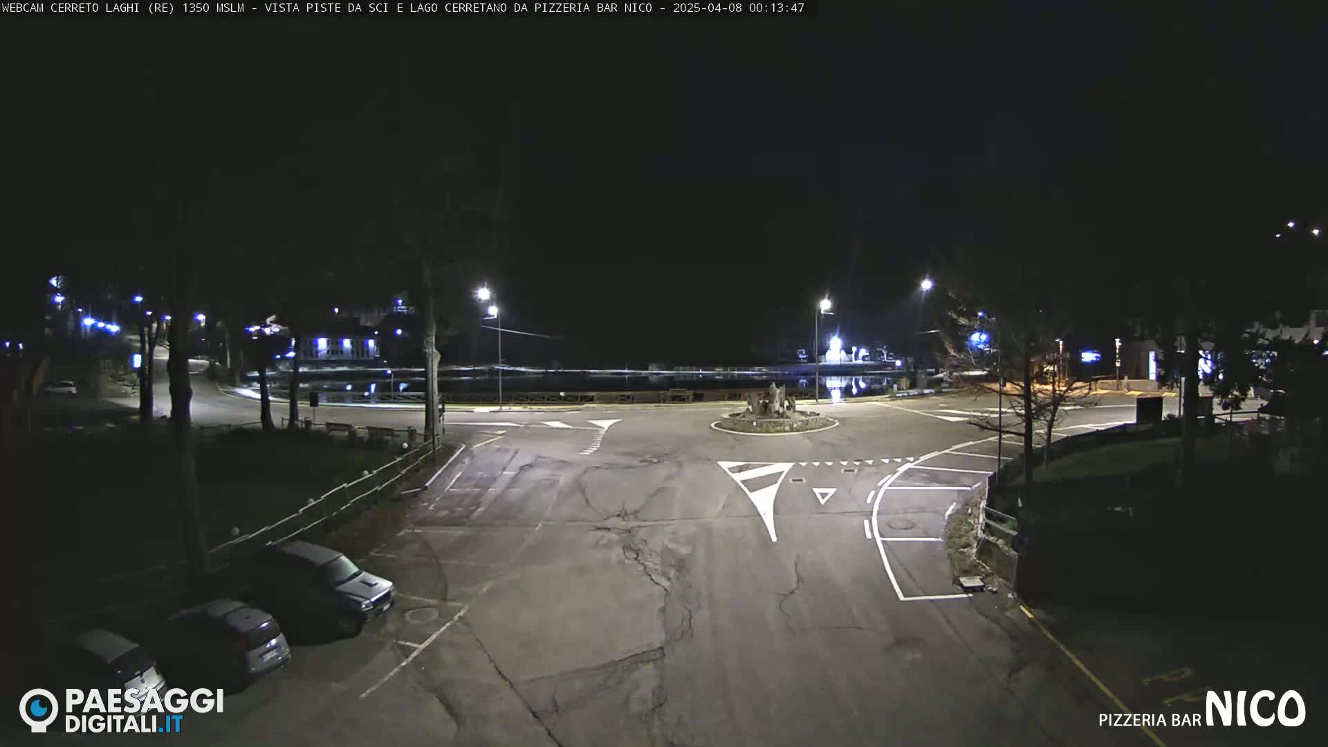 A nighttime view of a mostly empty asphalt roadway intersection with some parked cars, illuminated by streetlights.