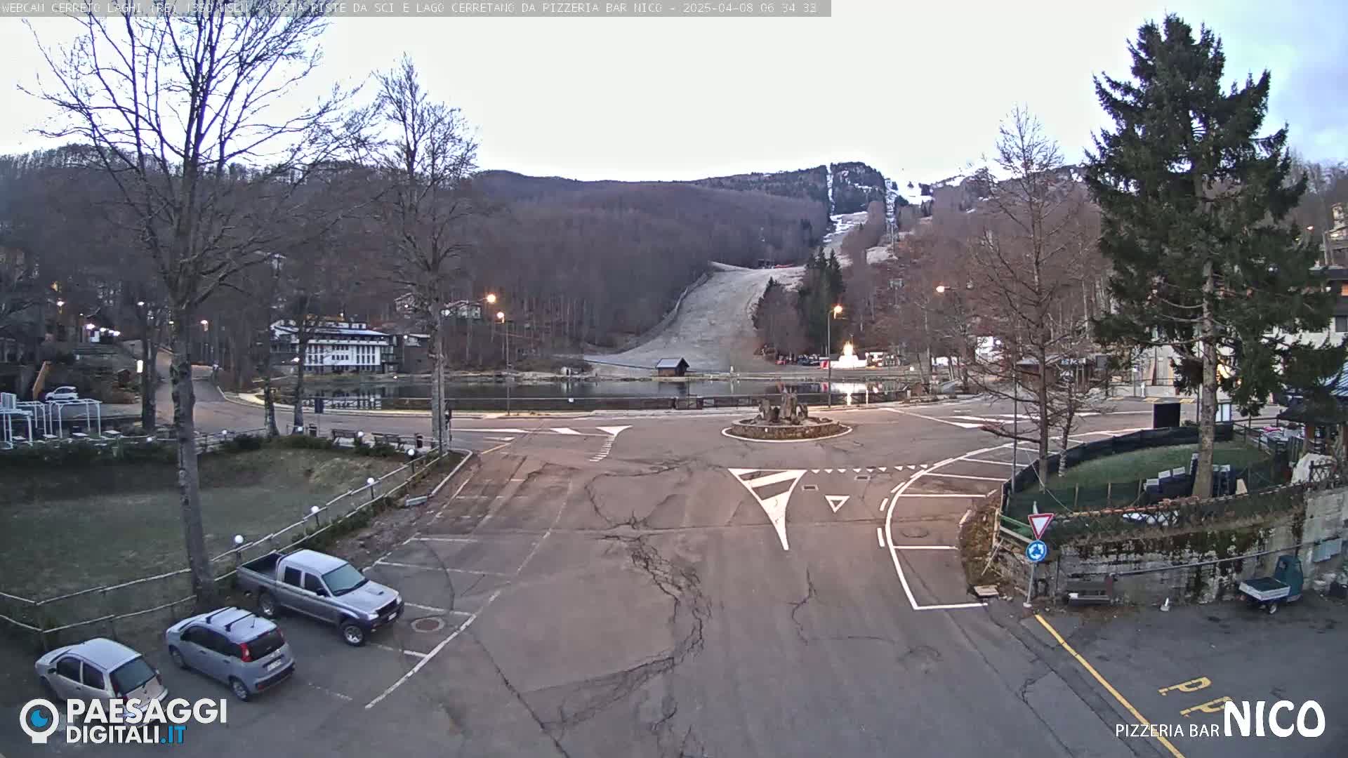 A mostly empty parking lot and intersection are seen, with a few cars parked, under an overcast sky, near a lake and ski slope in a wooded area.