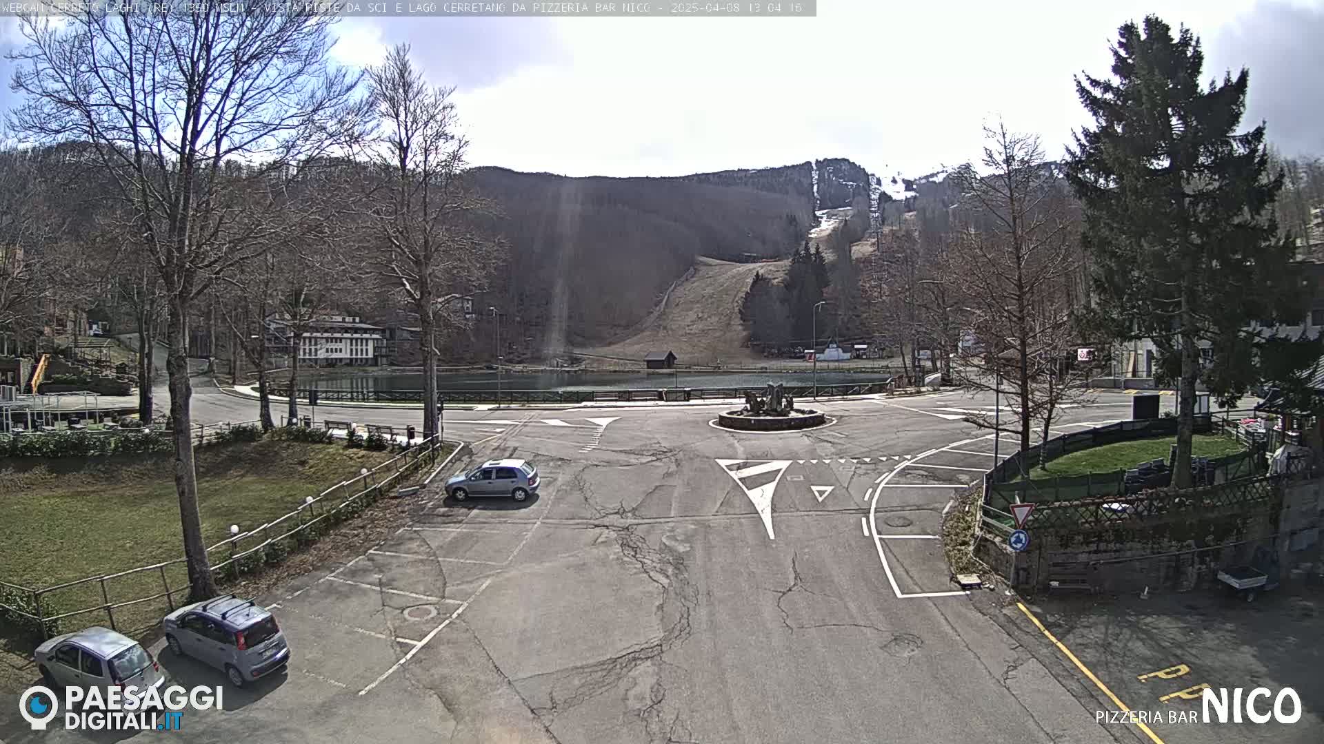A mostly sunny day reveals a parking lot and roundabout near a small lake and ski slope, surrounded by bare trees and buildings.