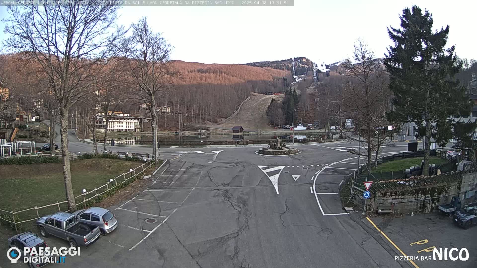 A mostly empty parking lot and roundabout are situated in front of a small town nestled beside a lake and ski slope, all under a clear, bright sky.
