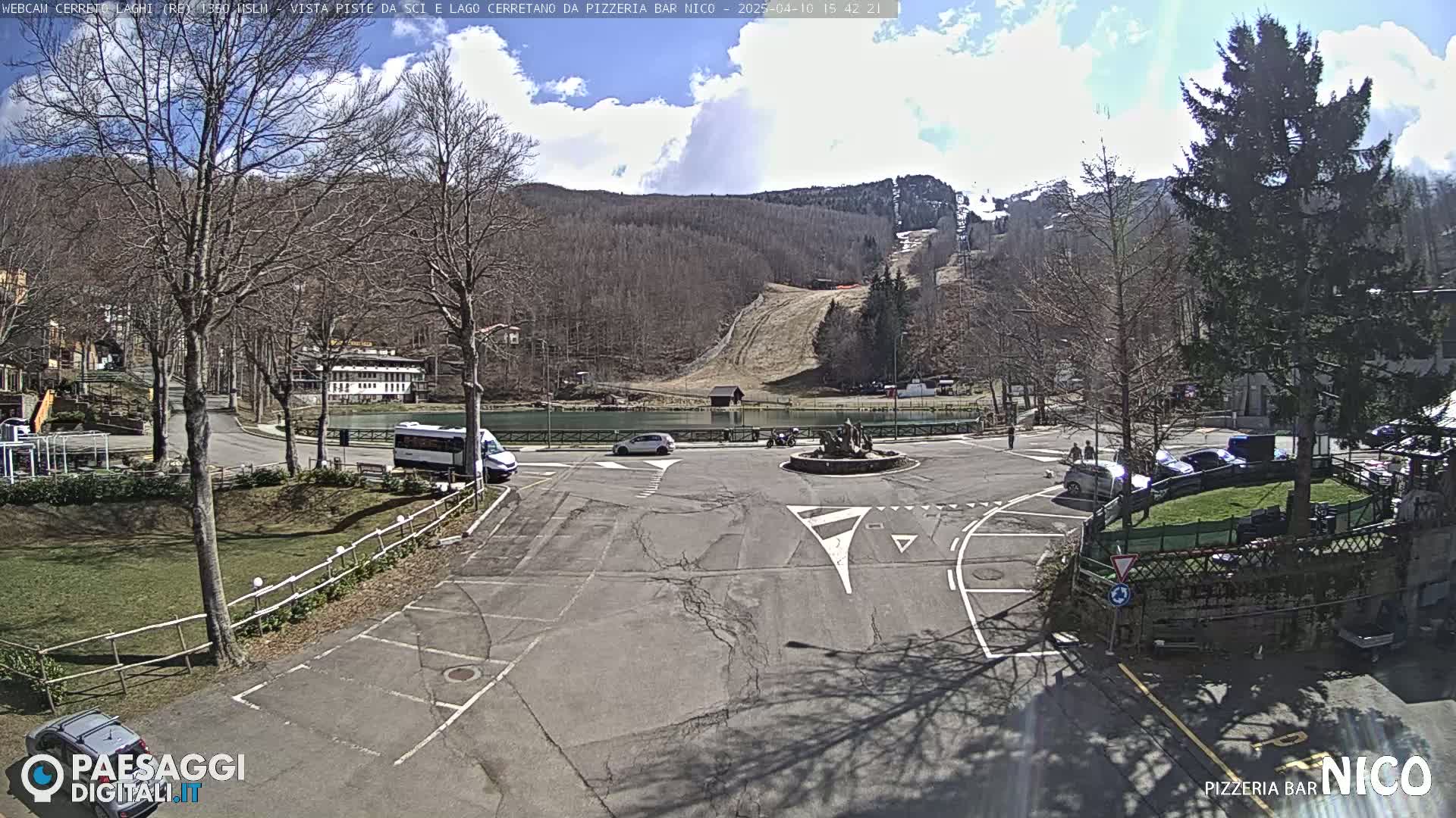 A parking lot and surrounding area, with a small lake and ski slope visible in the background, under a partly cloudy sky.