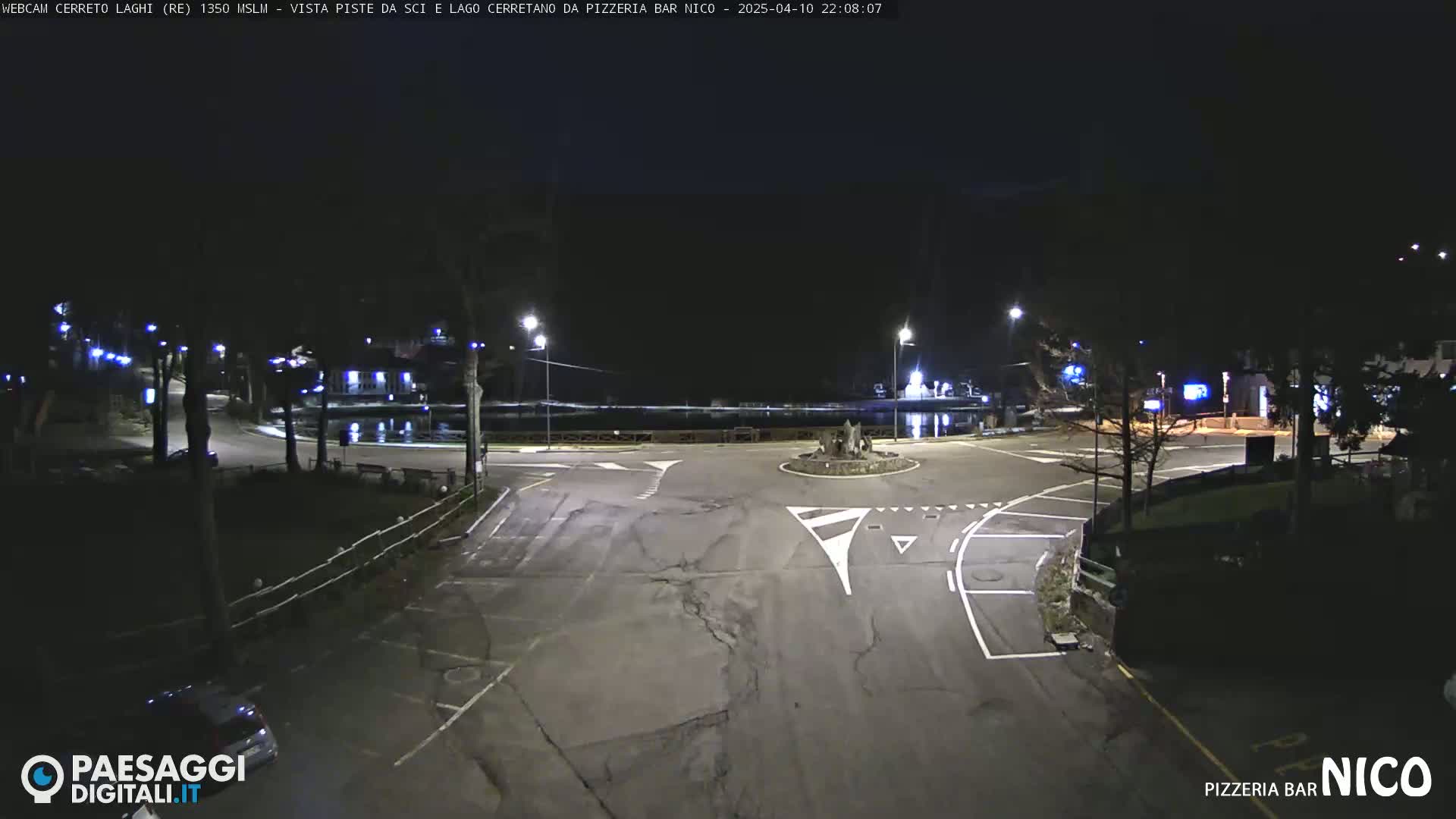 A nighttime view of a mostly empty roundabout and parking lot next to a body of water, illuminated by streetlights.