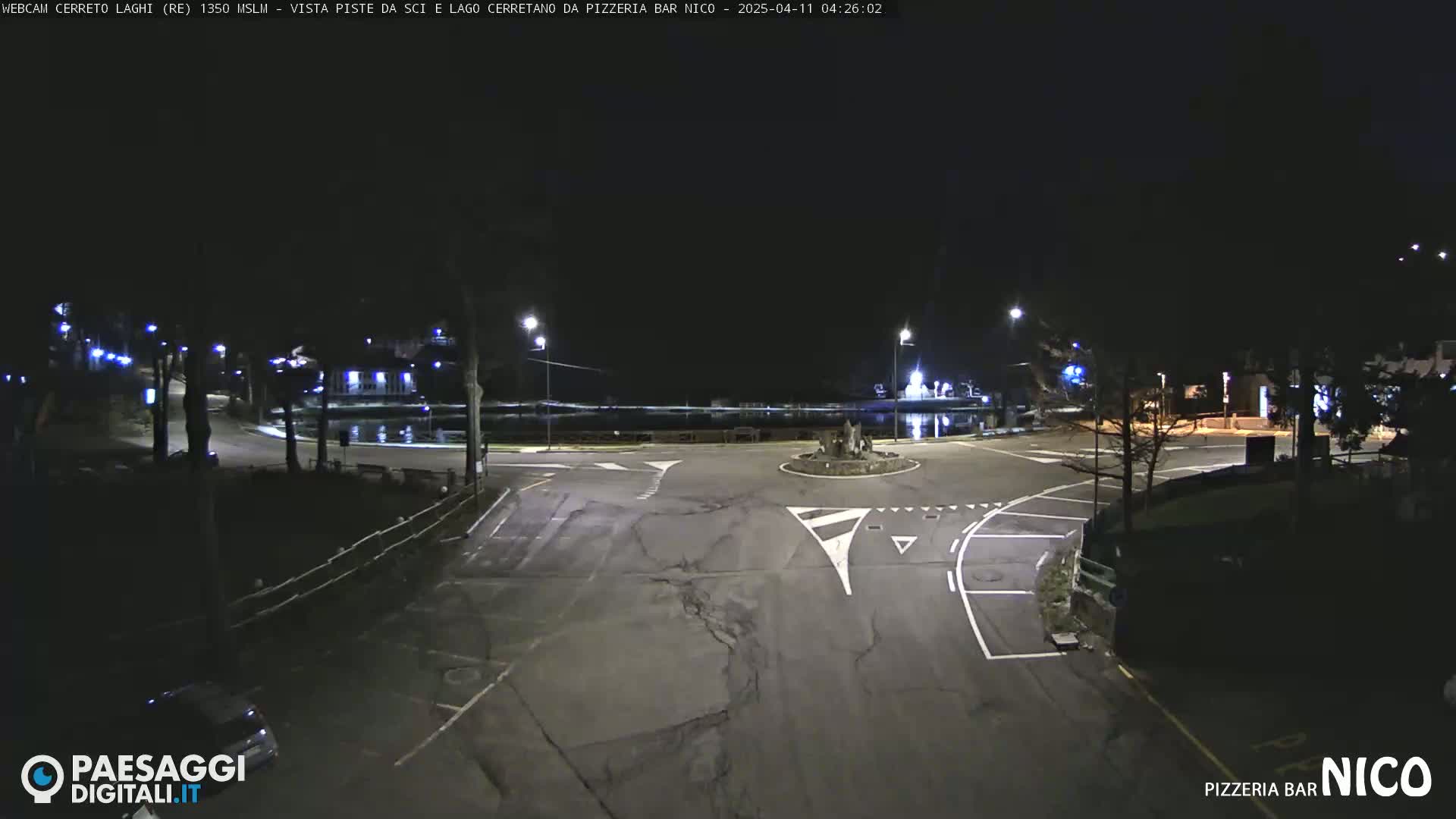 A nighttime view of a dark, empty roundabout near a body of water, illuminated by streetlights.