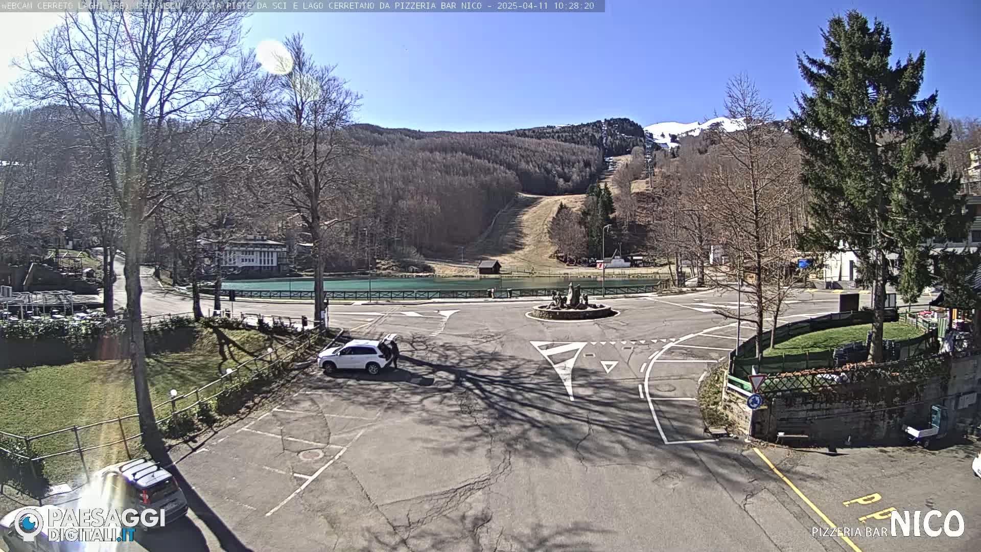 A sunny day reveals a parking lot and roundabout near a lake and ski slope in a wooded area.