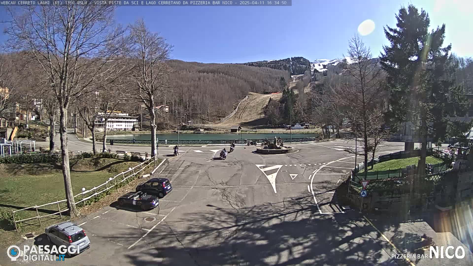 A sunny day reveals a parking lot with several cars, a small roundabout with a fountain, and a background of a ski slope and a lake, surrounded by bare trees and a snow-capped mountain in the distance.