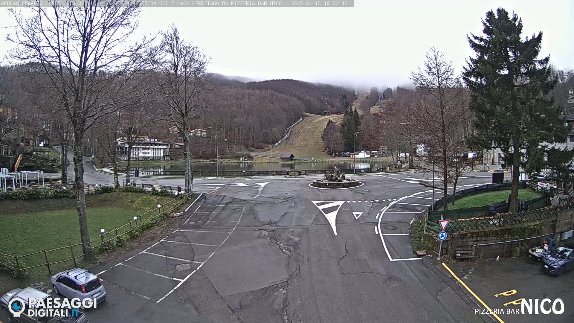 A mostly empty parking lot and roadway in a mountain town with a small pond, a ski slope in the background, and overcast weather.