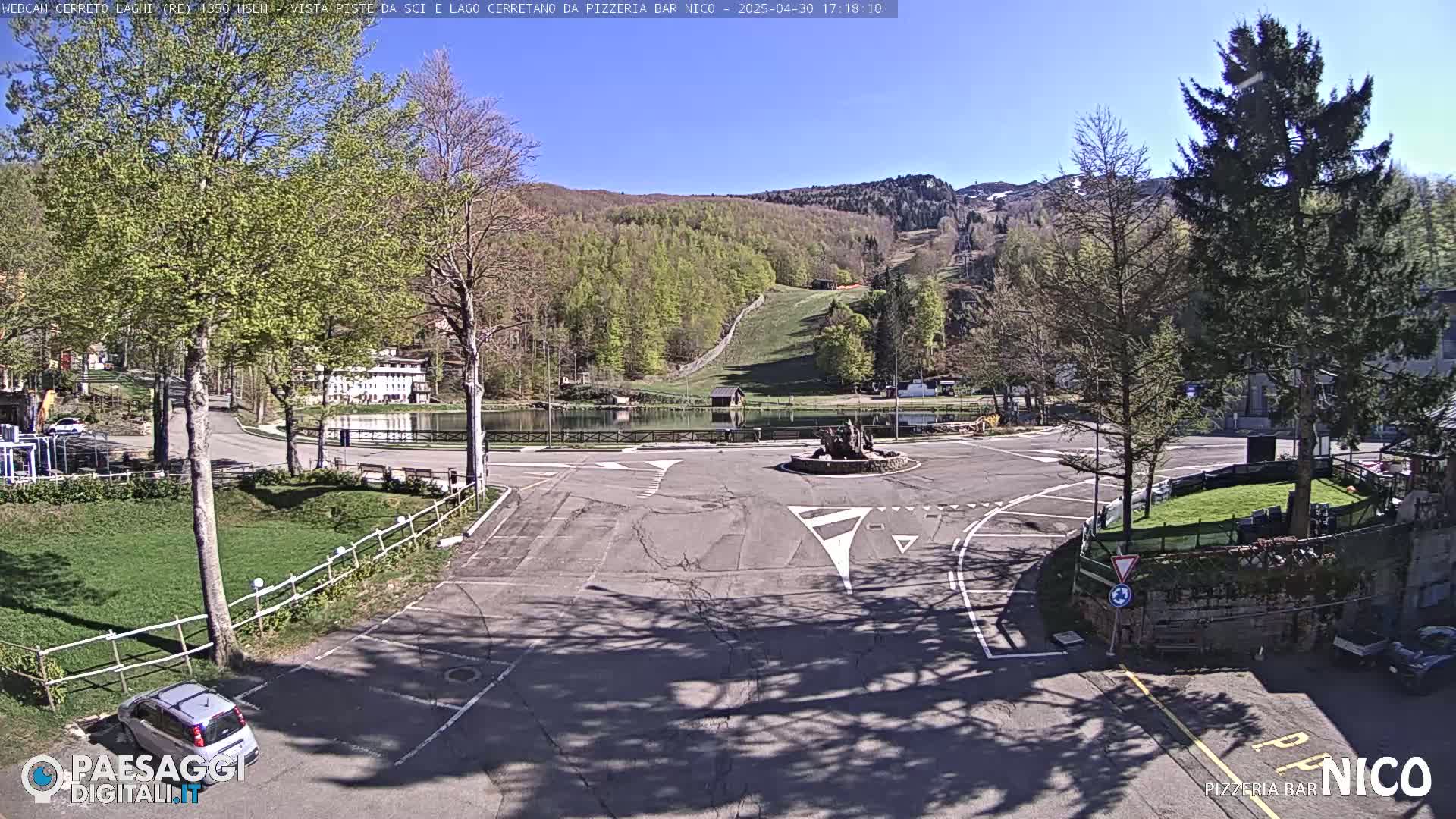 A sunny day reveals a parking lot and roundabout near a small lake and ski slope, surrounded by trees and buildings.