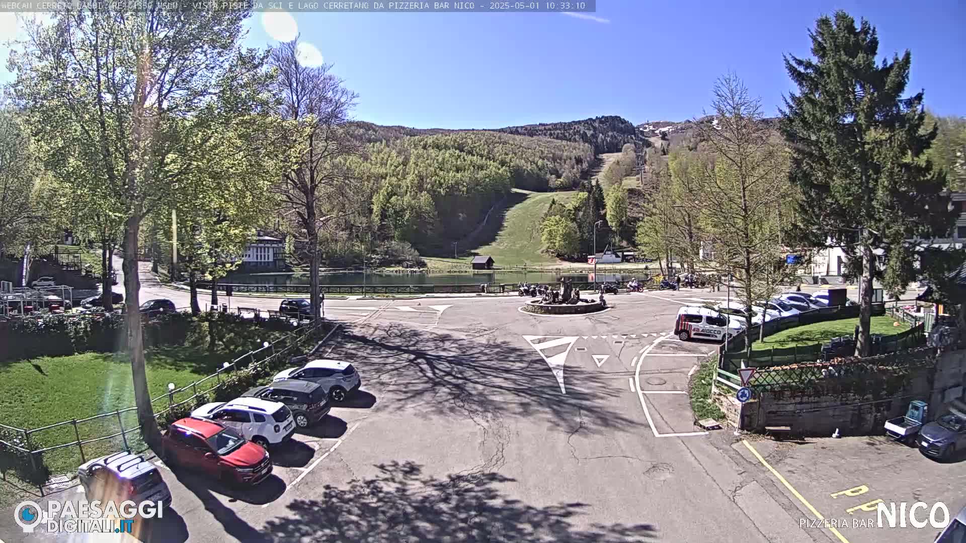 A sunny parking lot with several cars is situated near a small lake and a grassy hillside with a ski slope visible in the background.